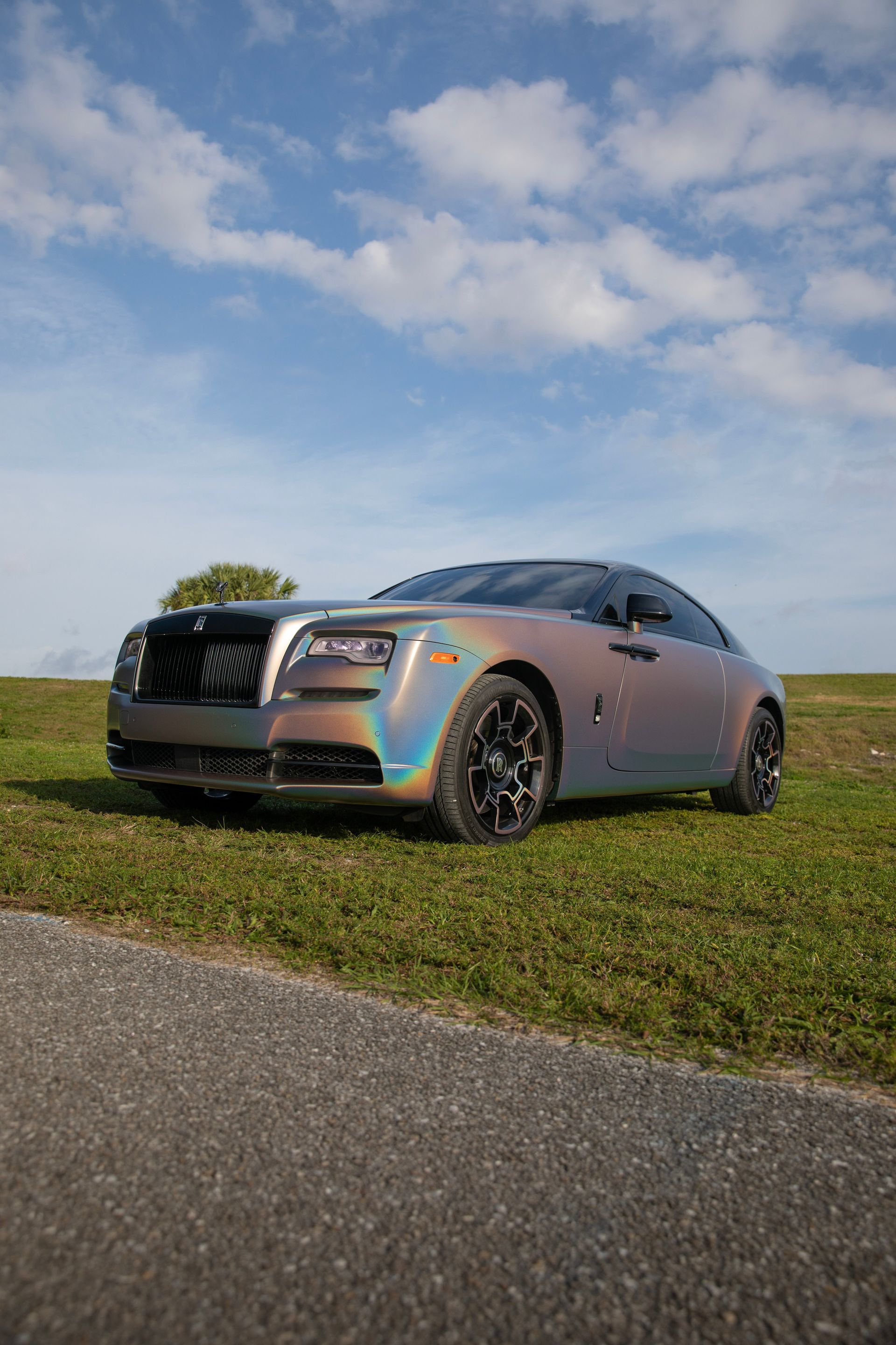 Iridescent Rolls-Royce coupe parked on green grass next to a road, under a blue sky.