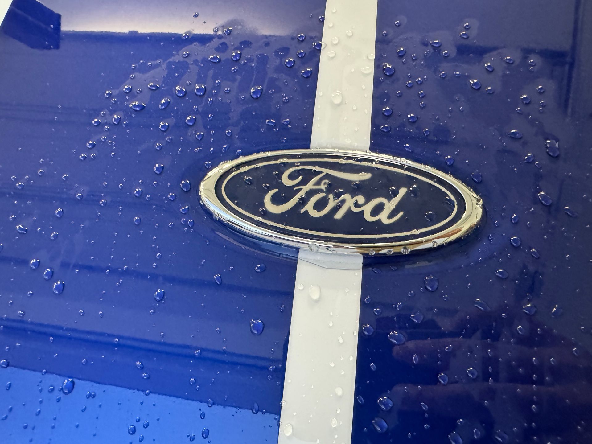 Blue Ford car hood with white stripe and logo, covered in water droplets.