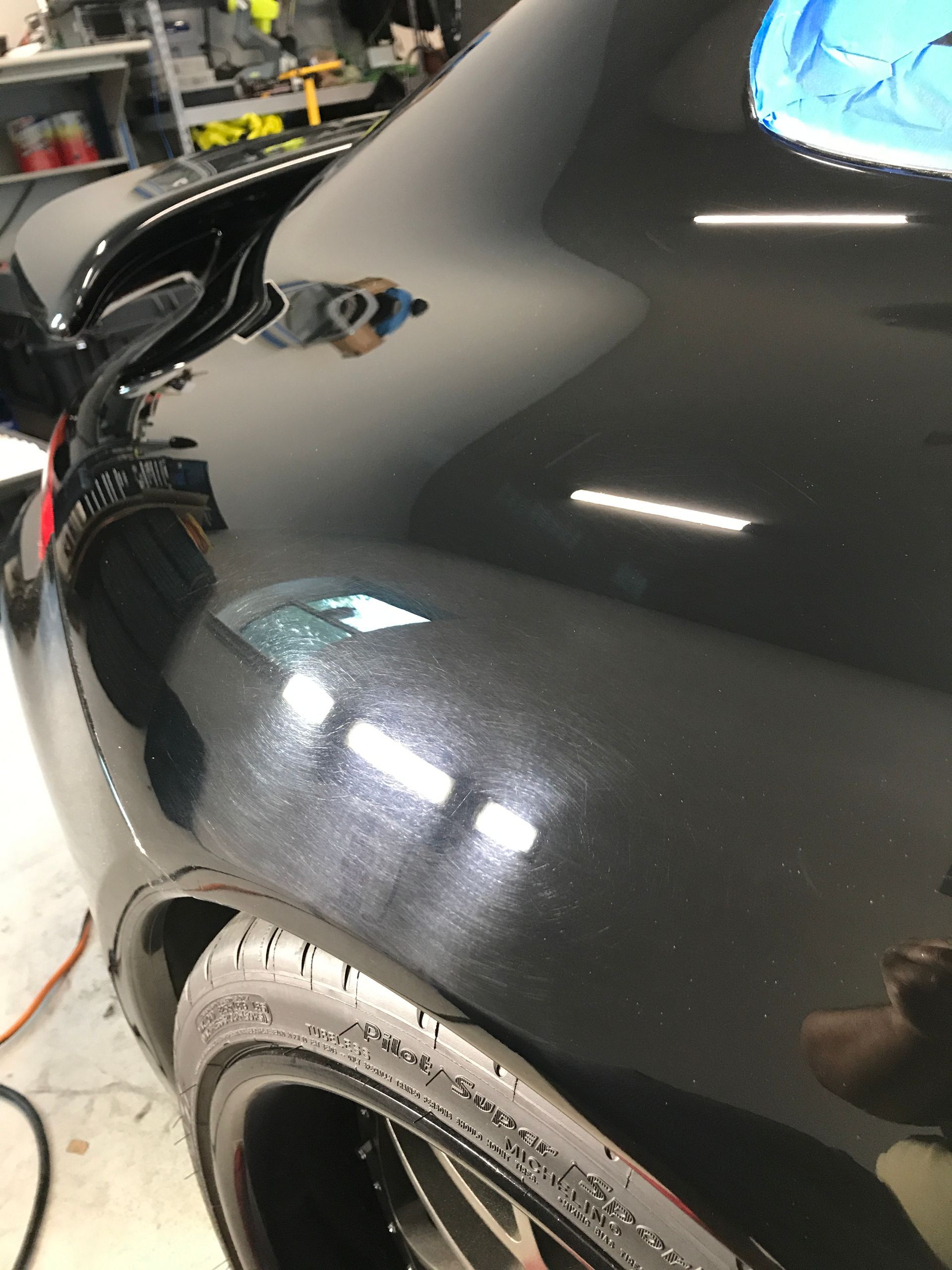 Black car's rear quarter panel showing visible swirls and reflections from overhead lights, likely in a detailing shop.