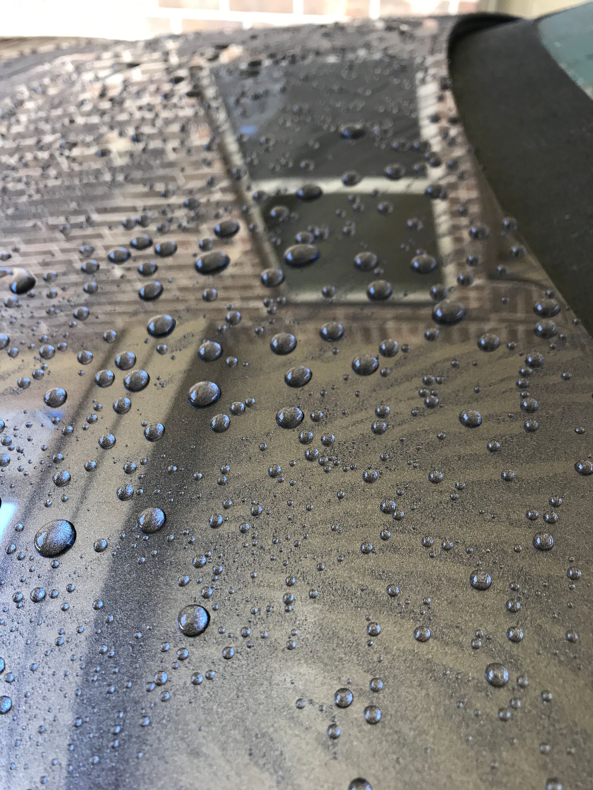 Water beads on a dark car surface, reflecting a window.