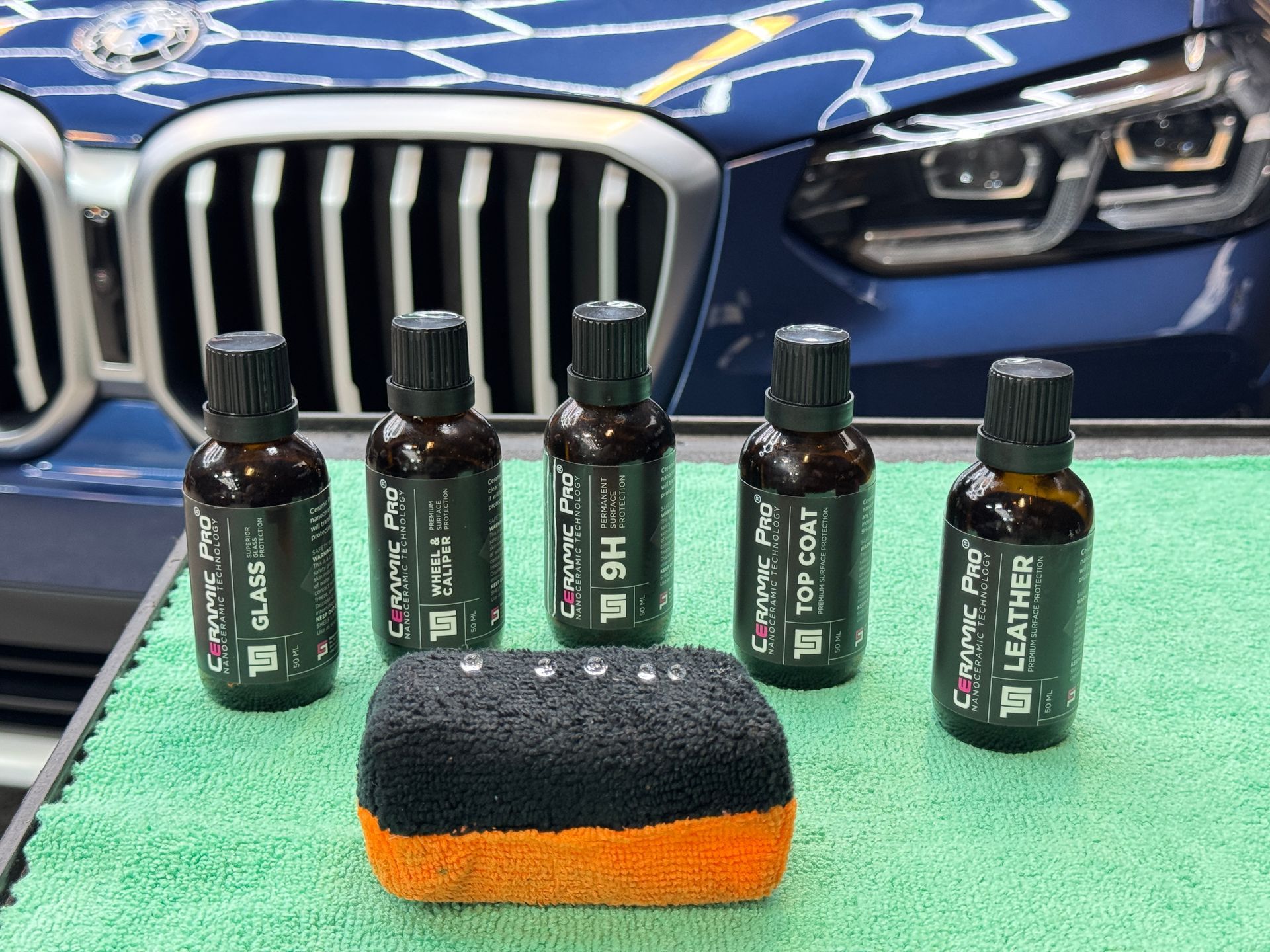 Car detailing products: Ceramic Pro bottles, applicator pad on a green towel in front of a blue BMW.