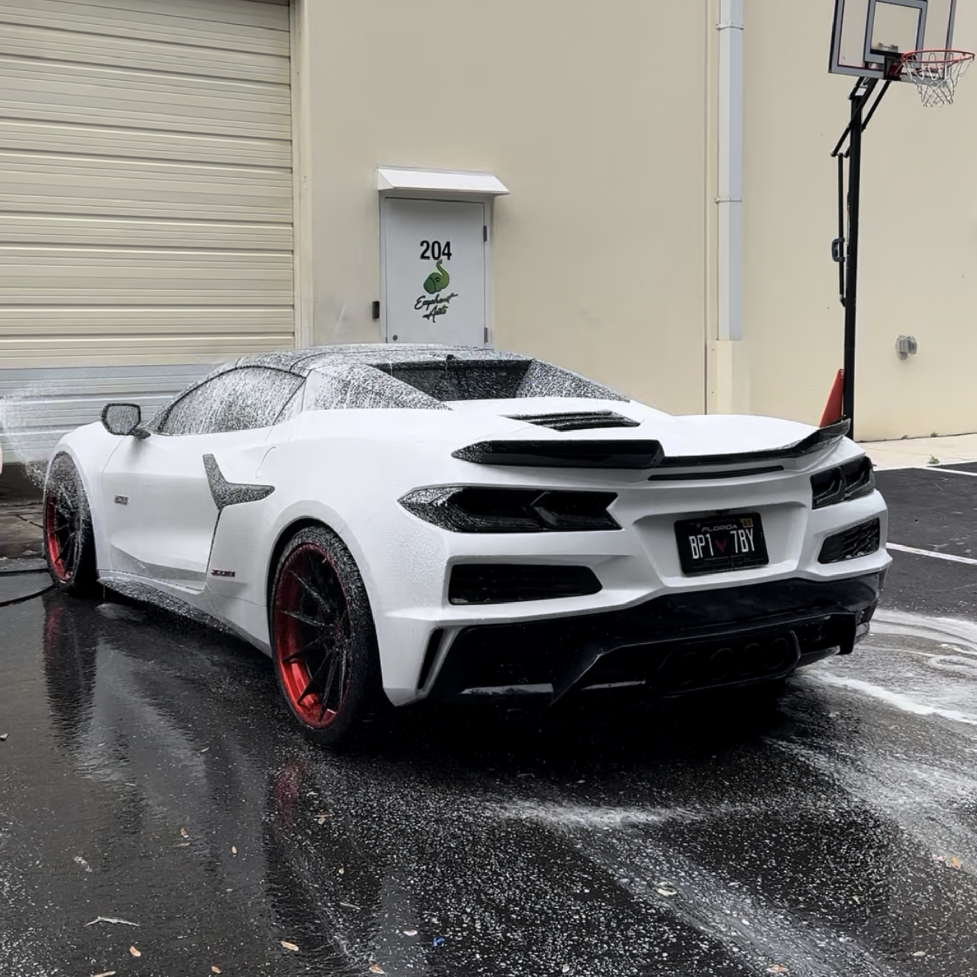 White sports car being washed; red rims, outdoor setting, building with basketball hoop visible.