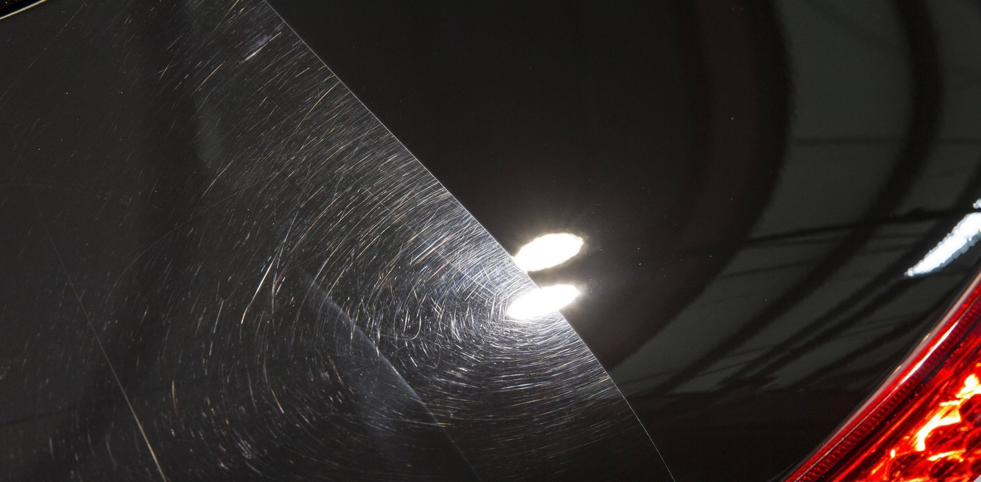 Close-up of a scratched black car surface reflecting bright lights and a red taillight.