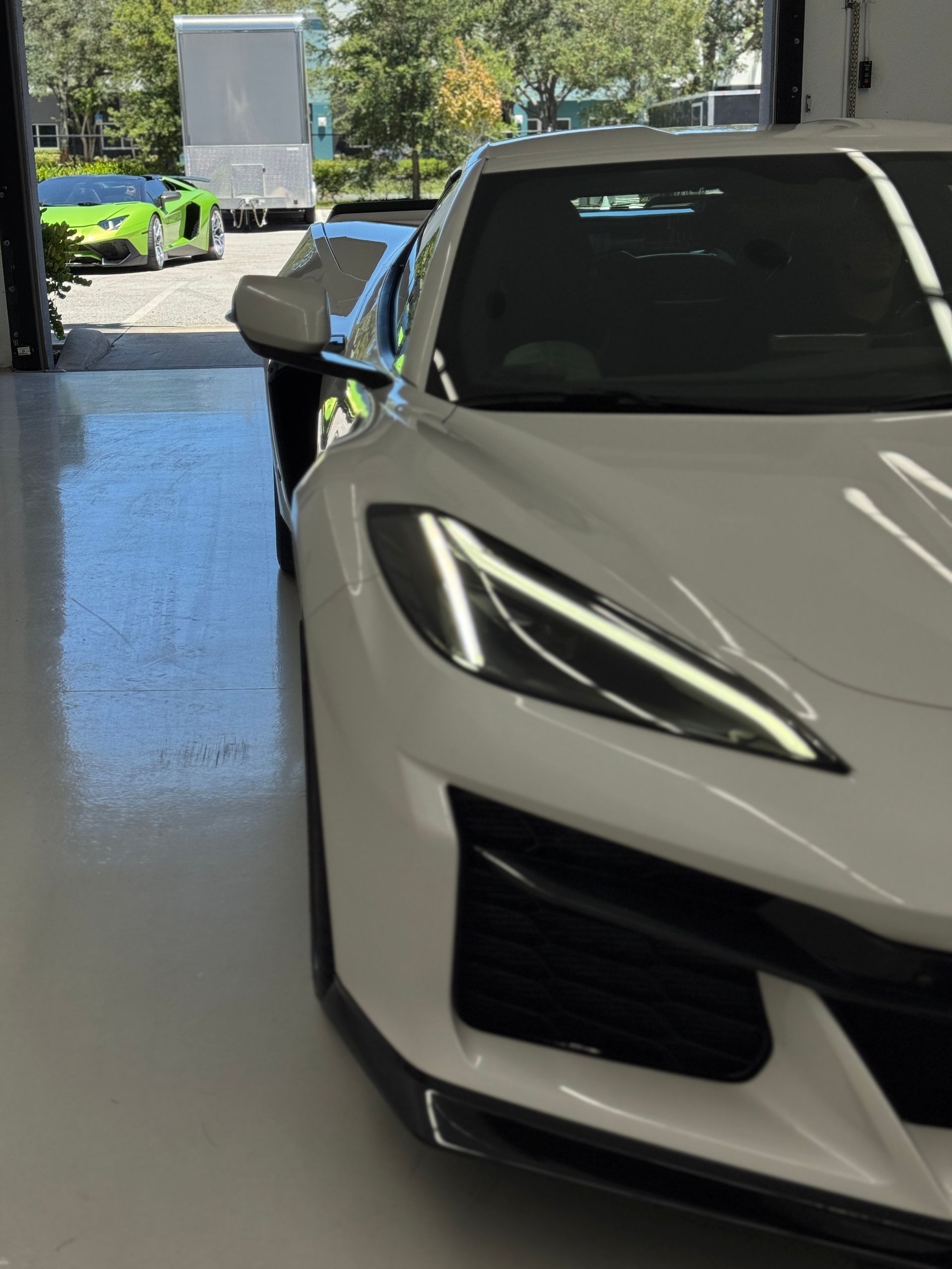White sports car in a garage, other cars in the background including a green Lamborghini.