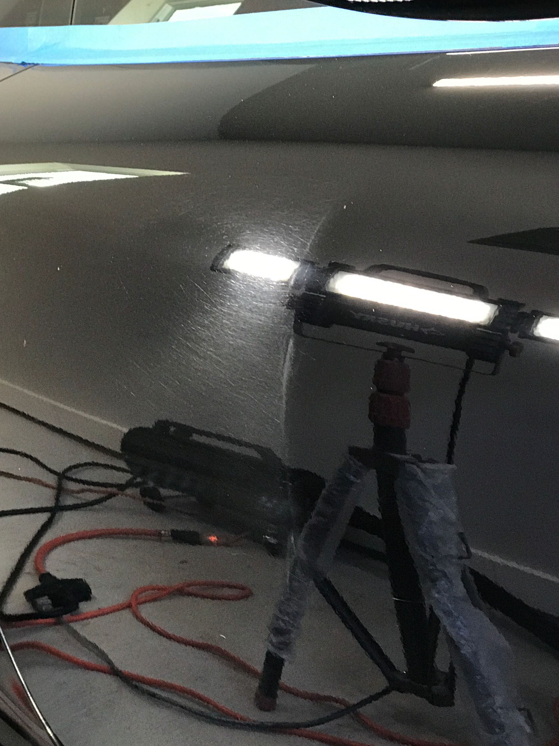 Black car hood illuminated by a bright work light on a tripod, wires visible.