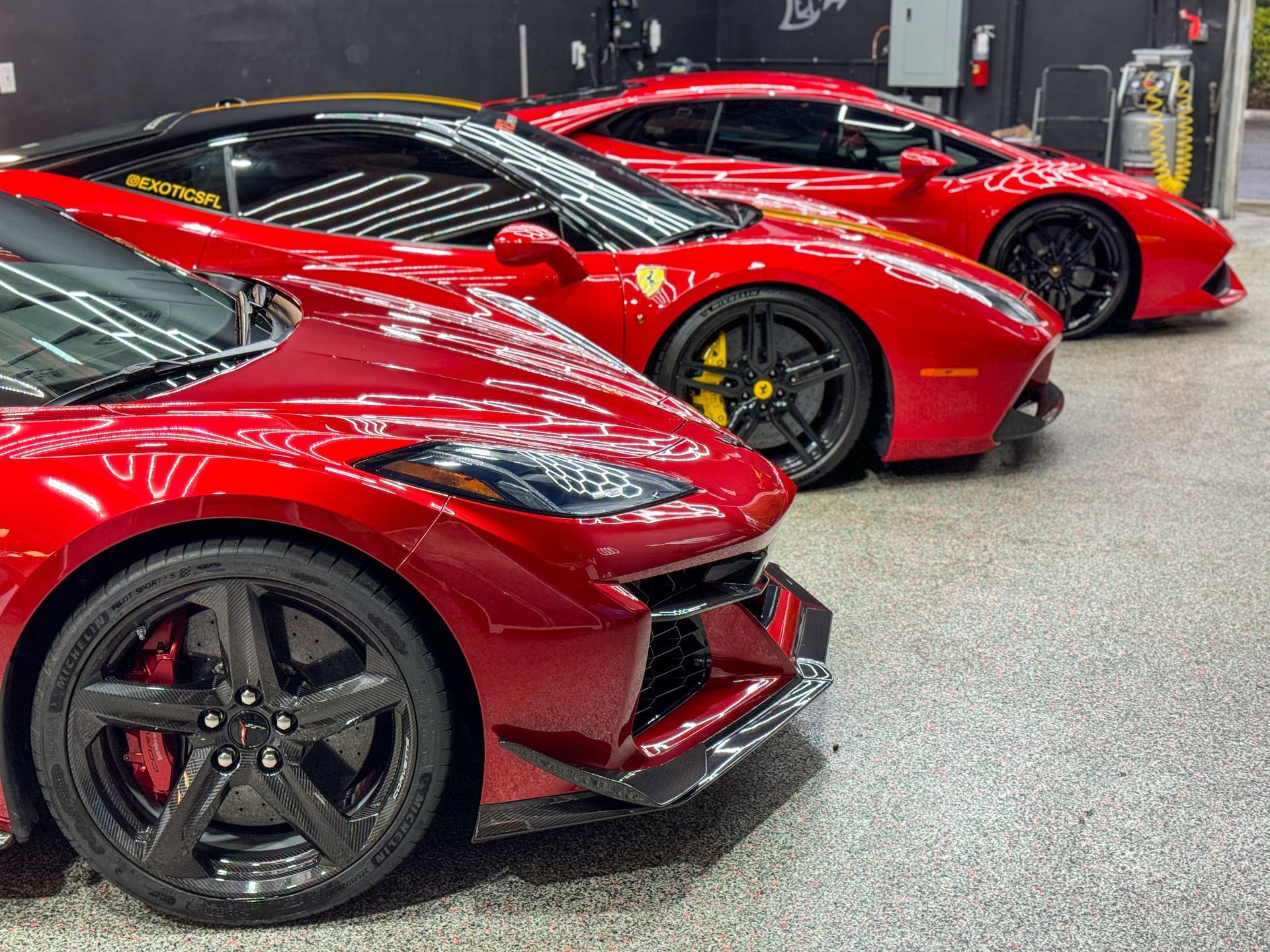 Three red sports cars parked in a garage, showcasing their details: a Corvette, a Ferrari, and another similar model.