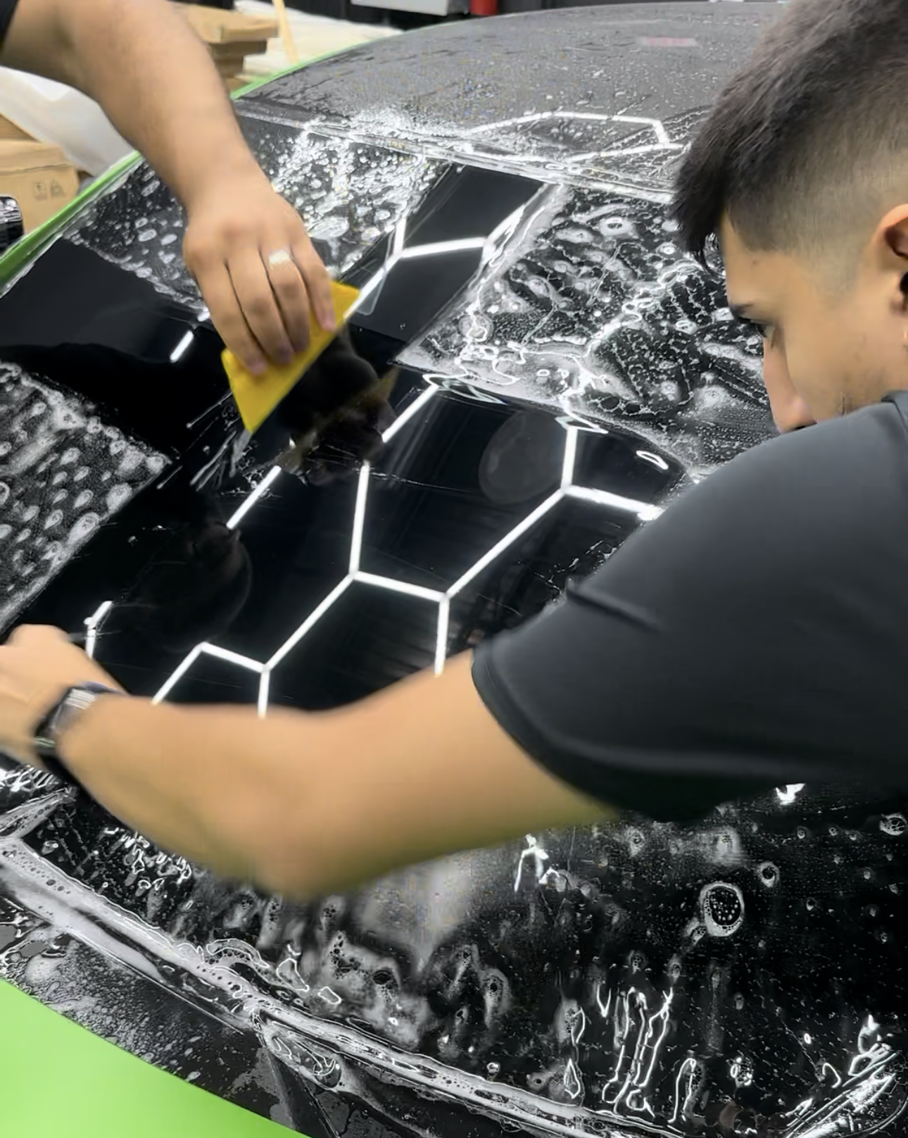 Two people installing black film on a car window, using a squeegee and soapy water.