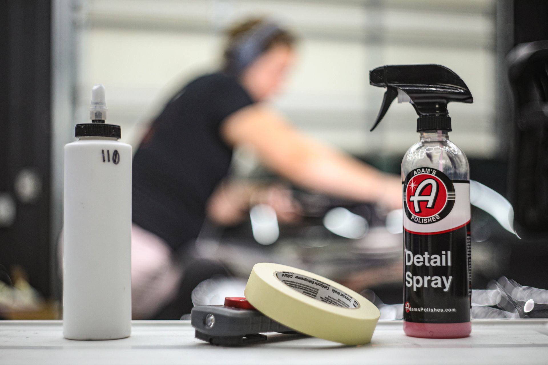 Car detailing supplies on table with blurred person working on vehicle in the background.