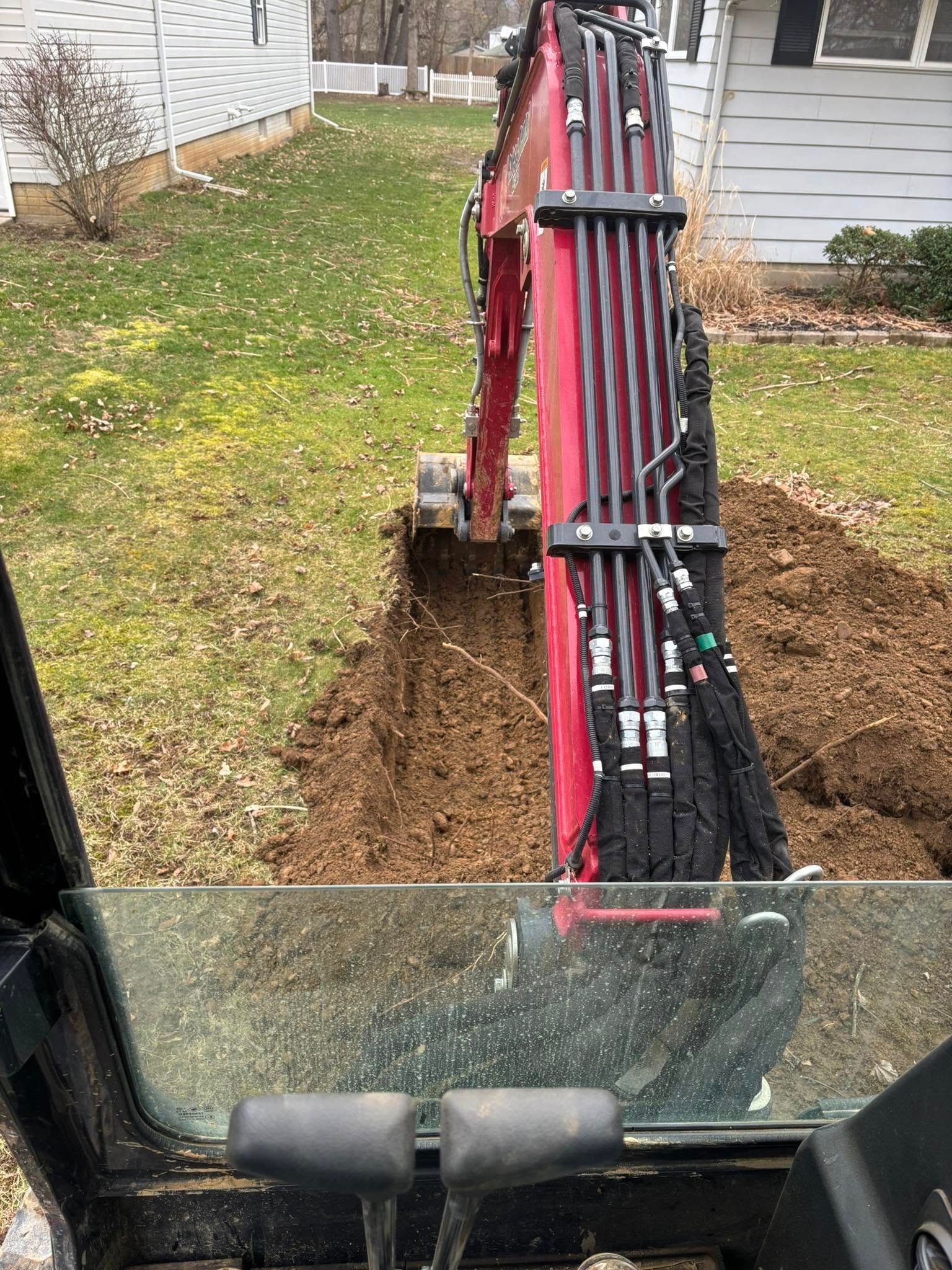 Excavator bucket digging a trench beside a house, with soil piled along the narrow excavation.