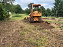 Orange bulldozer clearing muddy ground in a grassy field under cloudy skies