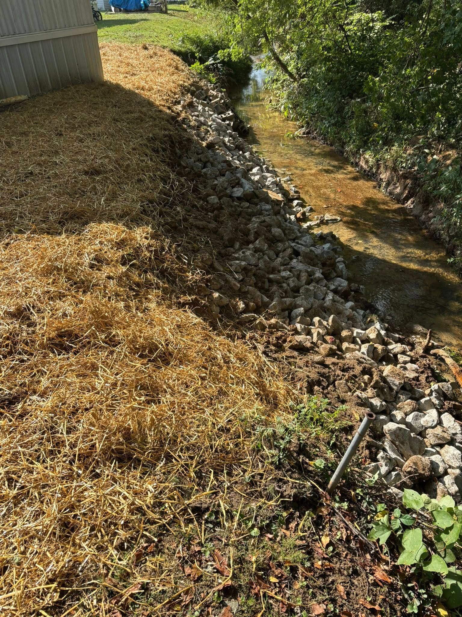 Narrow rocky drainage ditch beside a fence, lined with dry brown leaves and sparse greenery.