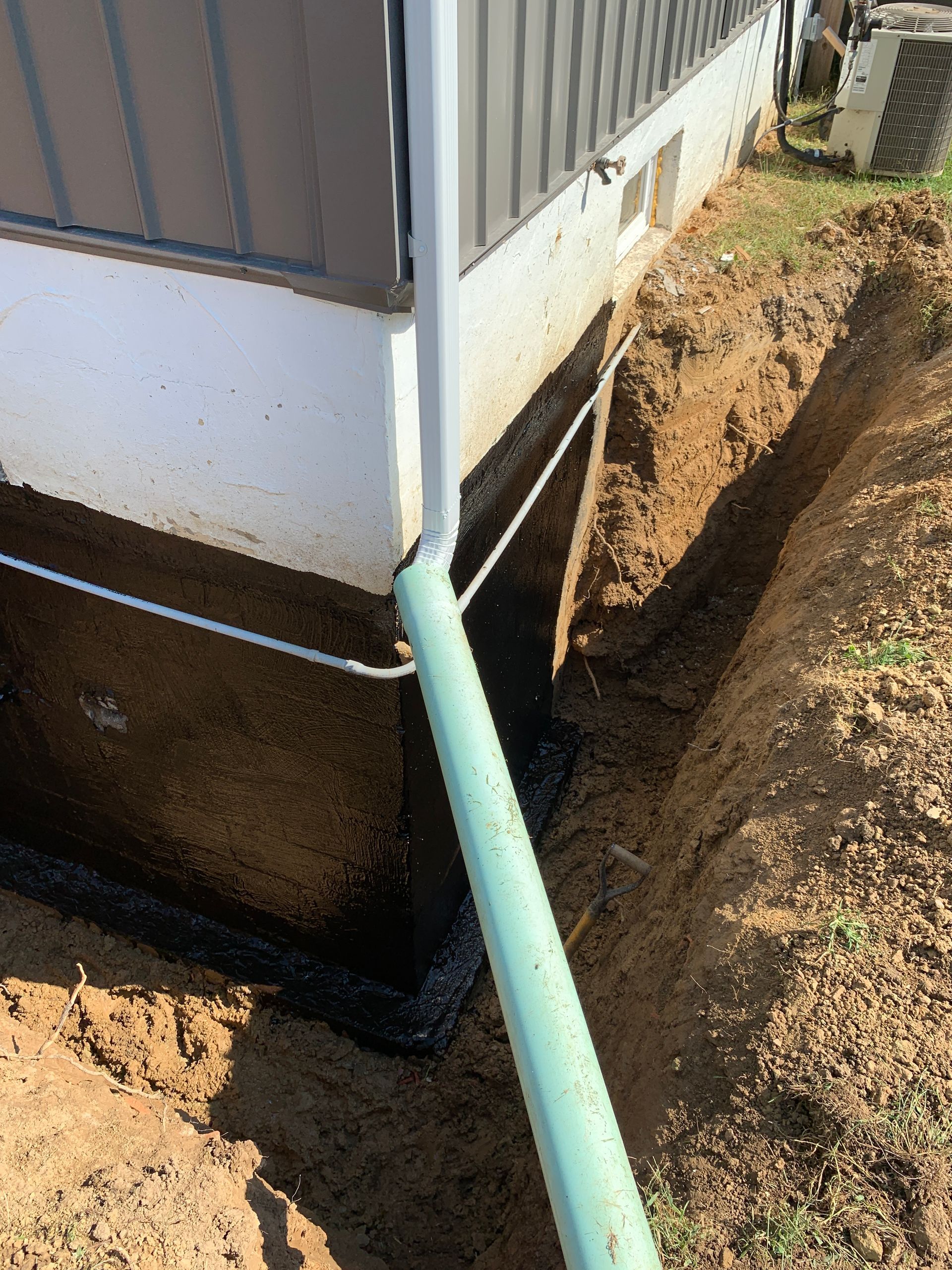 Excavated trench beside a building with a green downspout draining into black pipe and exposed foundation.