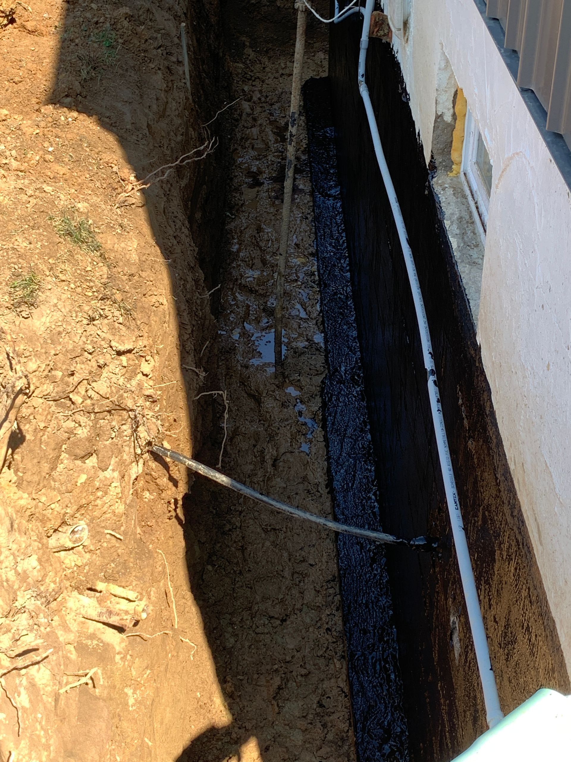 Narrow trench beside a building wall with exposed dirt and a blue pipe or cable running through it.
