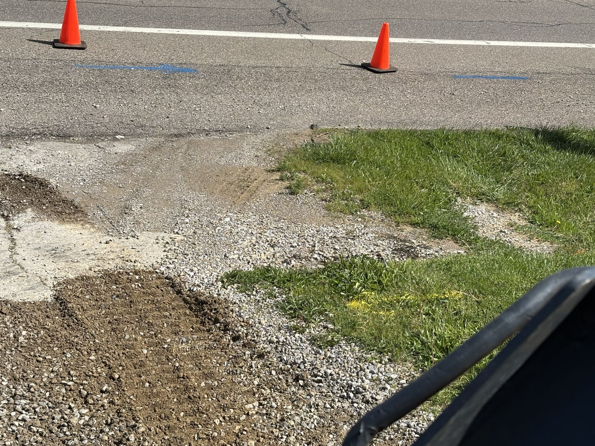 Paved road with pothole and gravel patch, marked by orange traffic cones near a grassy edge