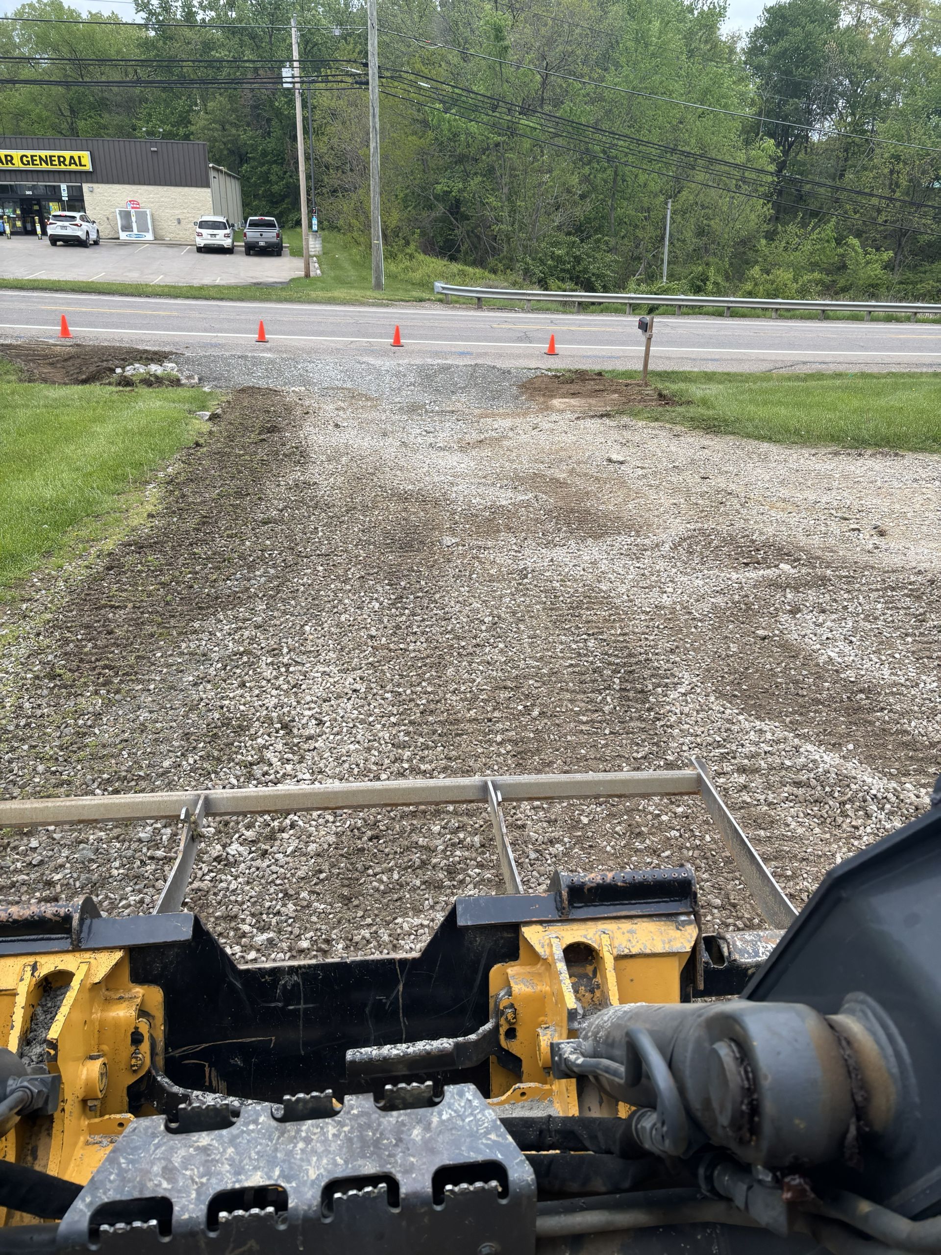View from a yellow excavator over a gravel road under construction, with traffic cones and trees ahead