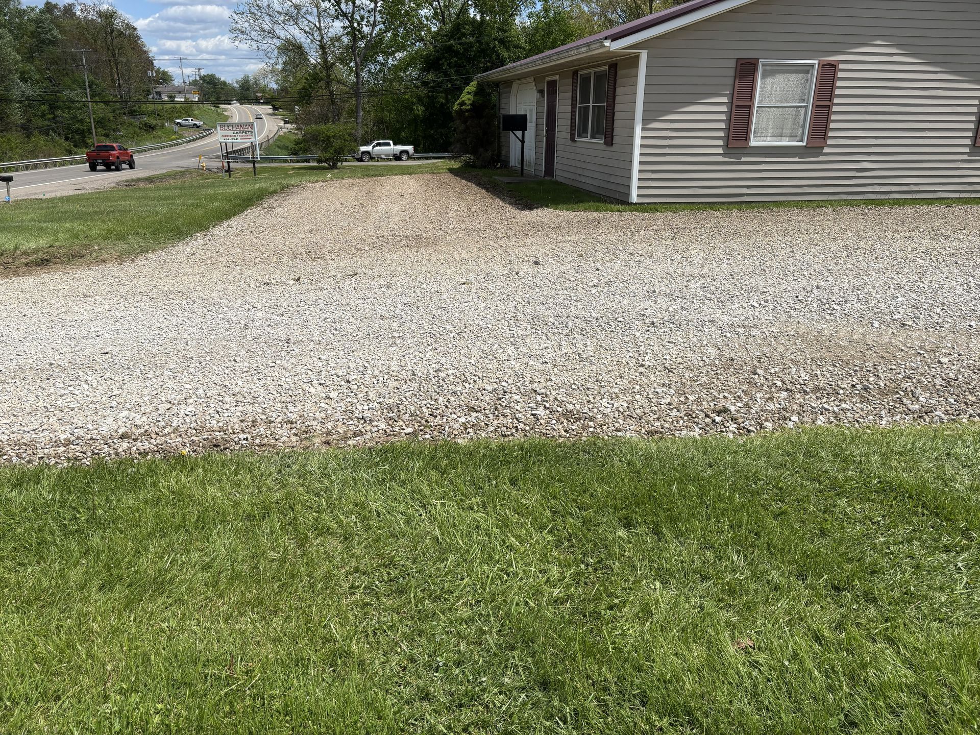 Gravel driveway beside a beige house, with a green lawn and trees in the background.