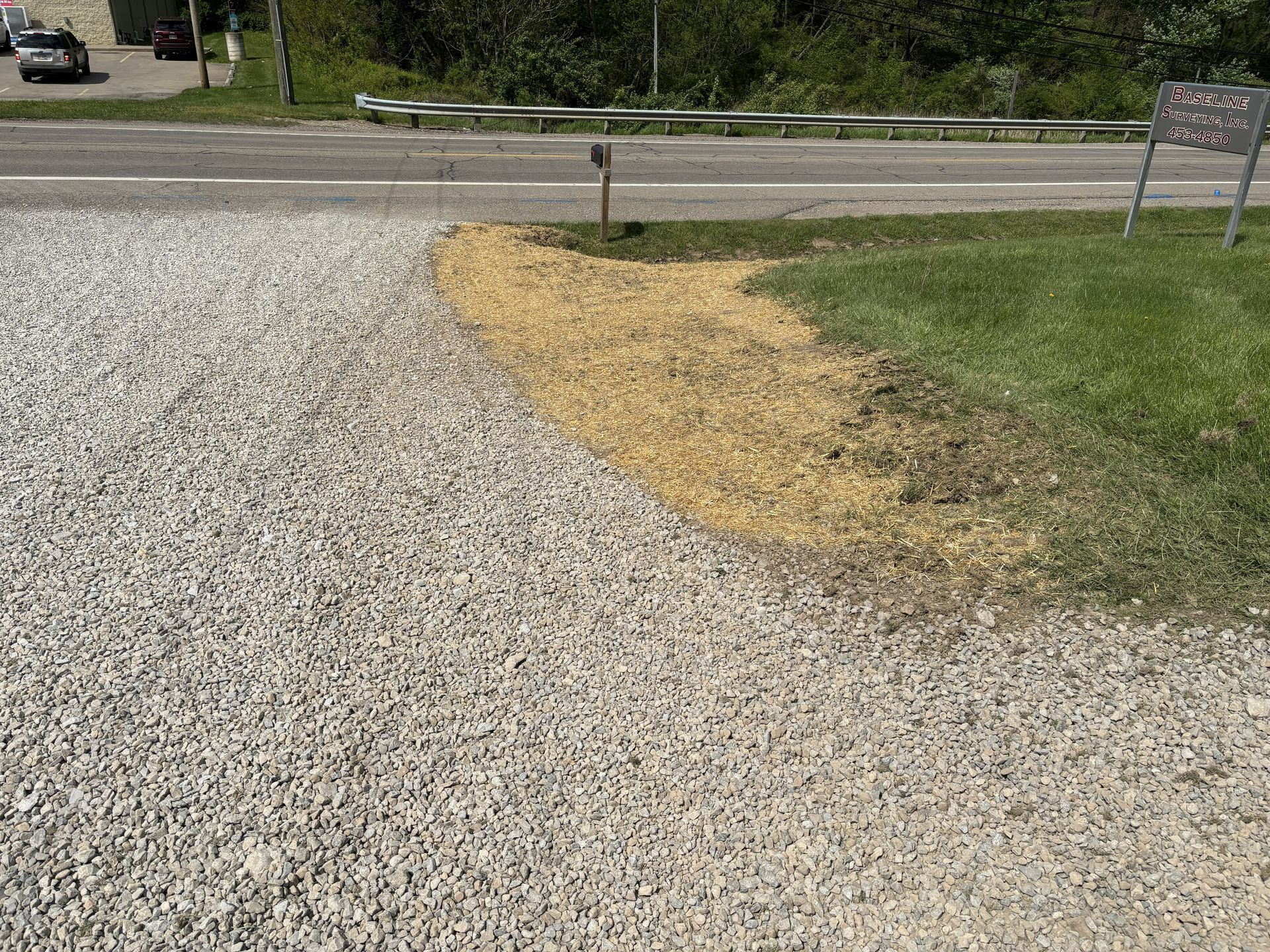 Gravel driveway curving toward a road, with a patch of yellowed grass and trees in the background.