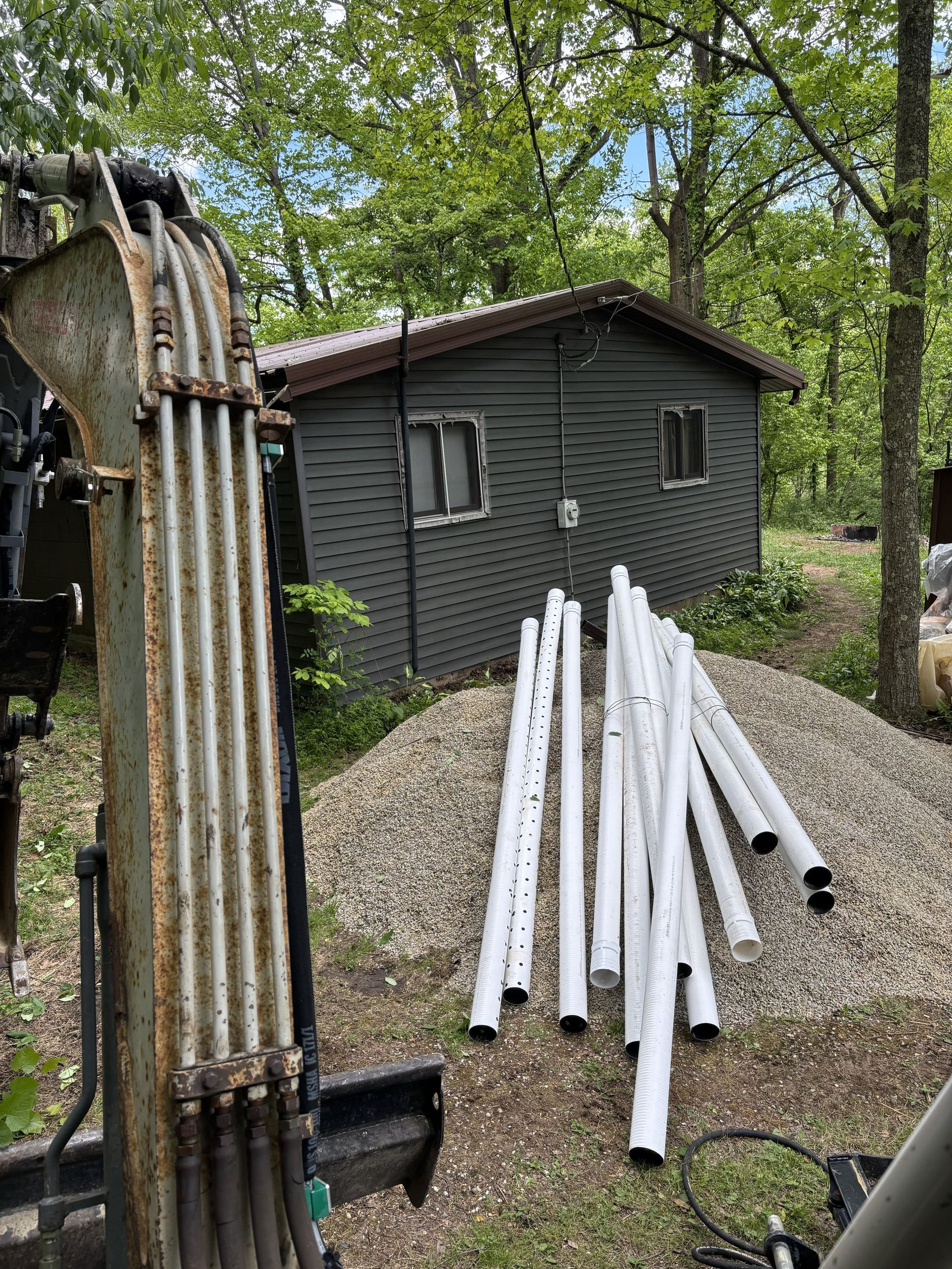 Gray shed beside trees with white PVC pipes stacked on gravel, next to an excavator arm