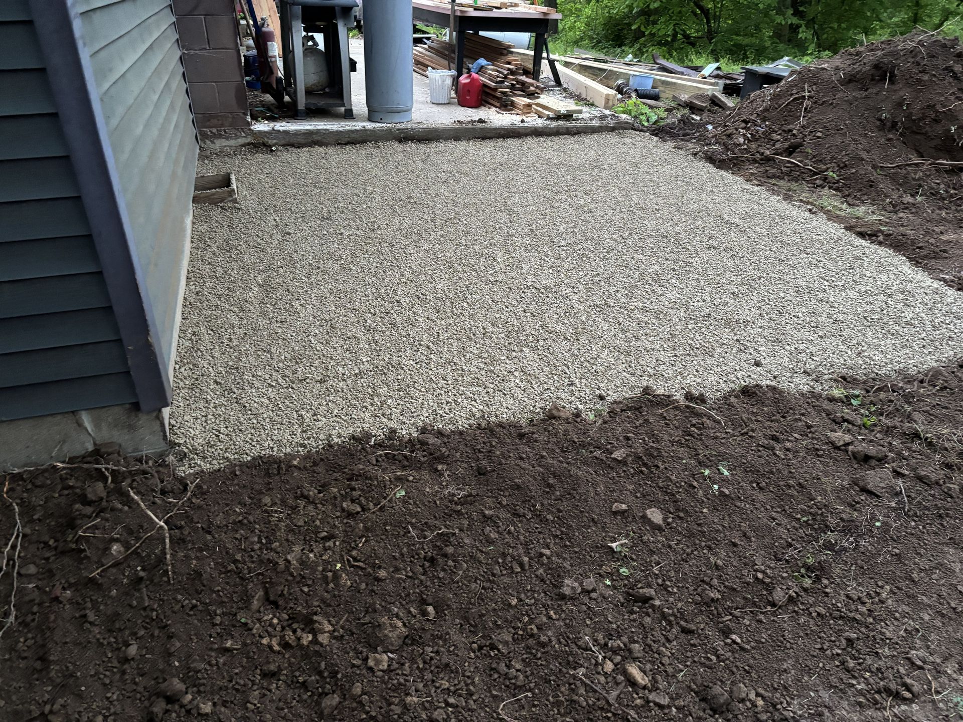 Gravel walkway beside a house, bordered by dark soil and a small patio area in the background