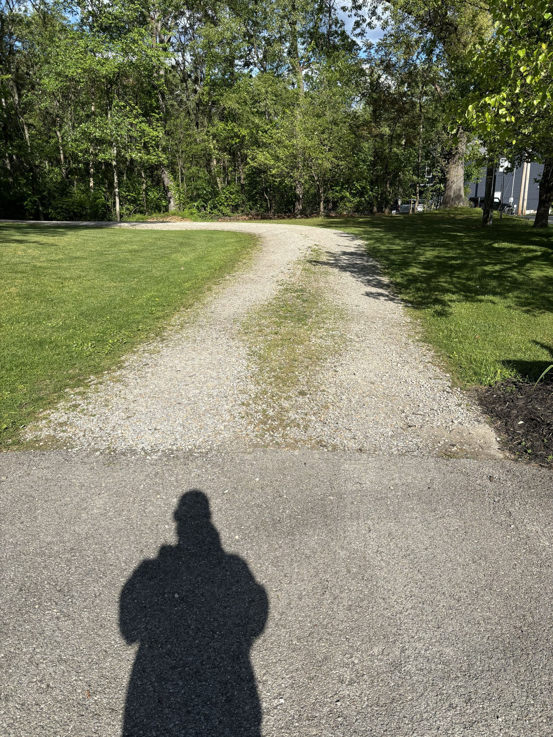 Gravel path leading through a sunny green park with trees; person’s shadow in the foreground