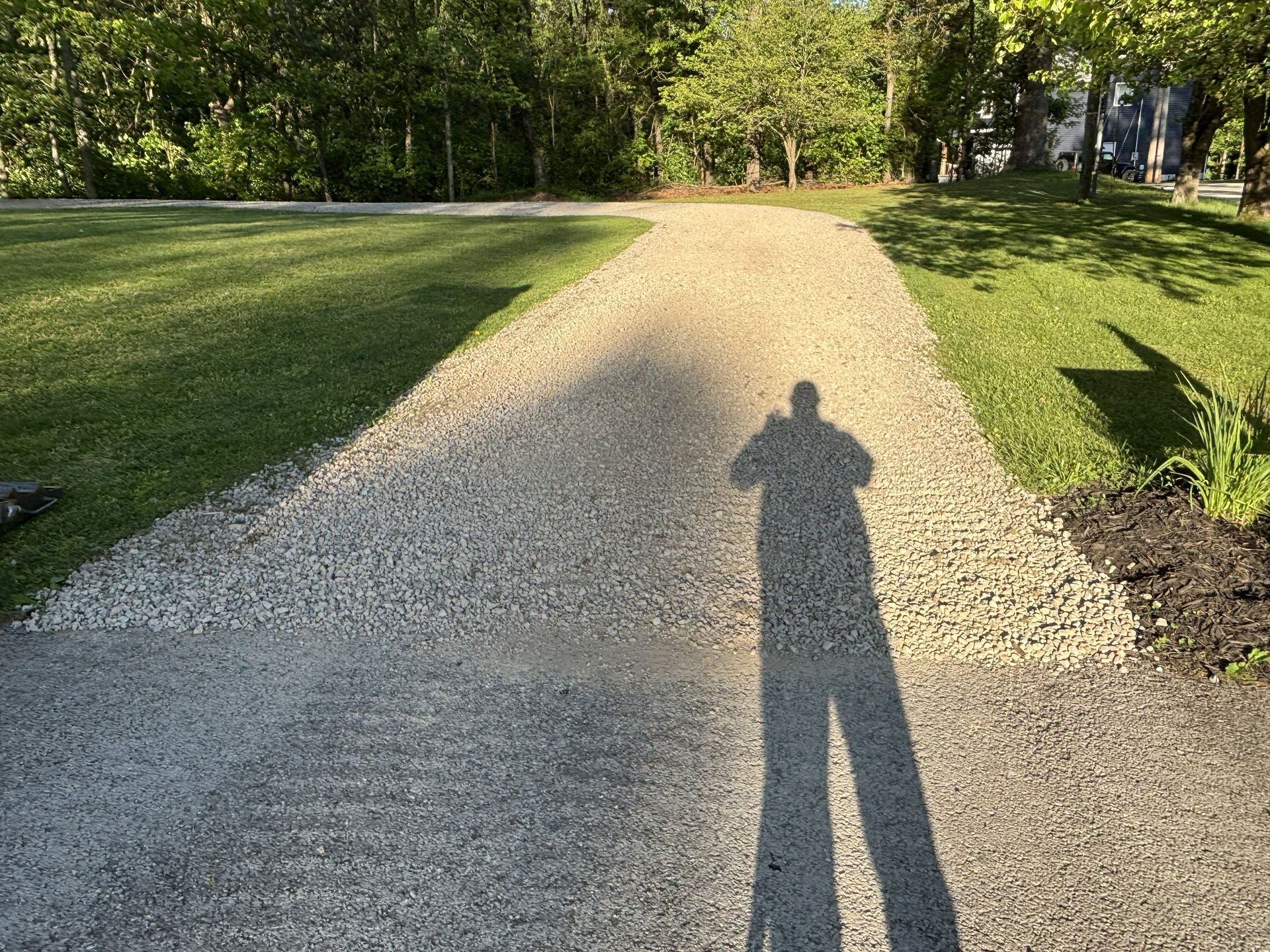 Sunlit gravel path through a green park, with a long shadow in the foreground.