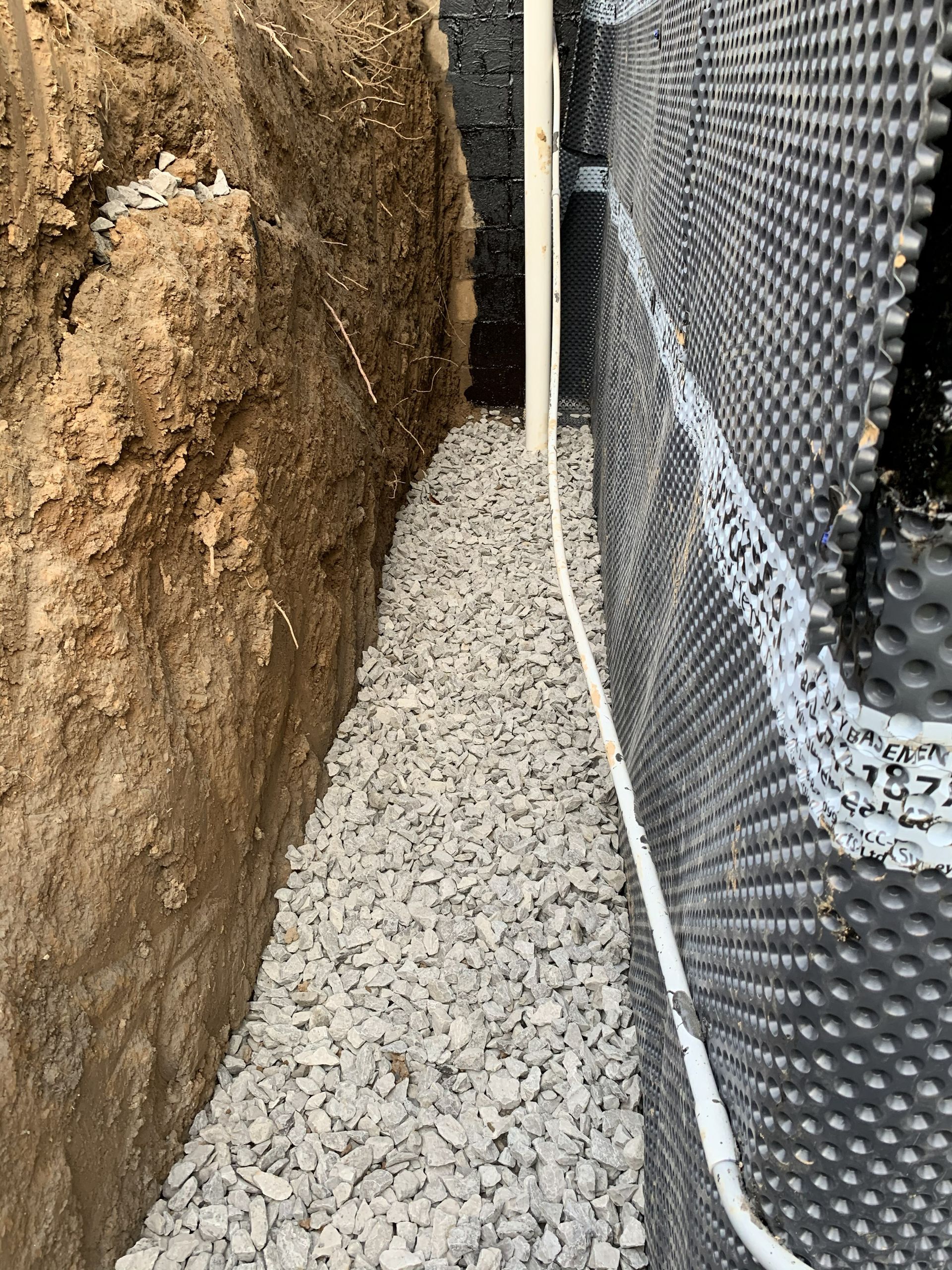 Narrow gravel trench beside a dirt wall and black waterproofed foundation