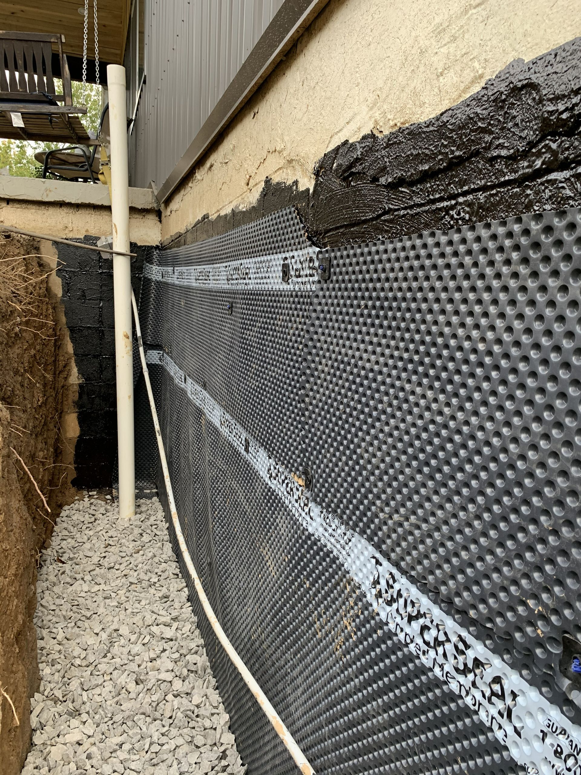 Foundation wall with black waterproof membrane and gravel beside a narrow outdoor passage