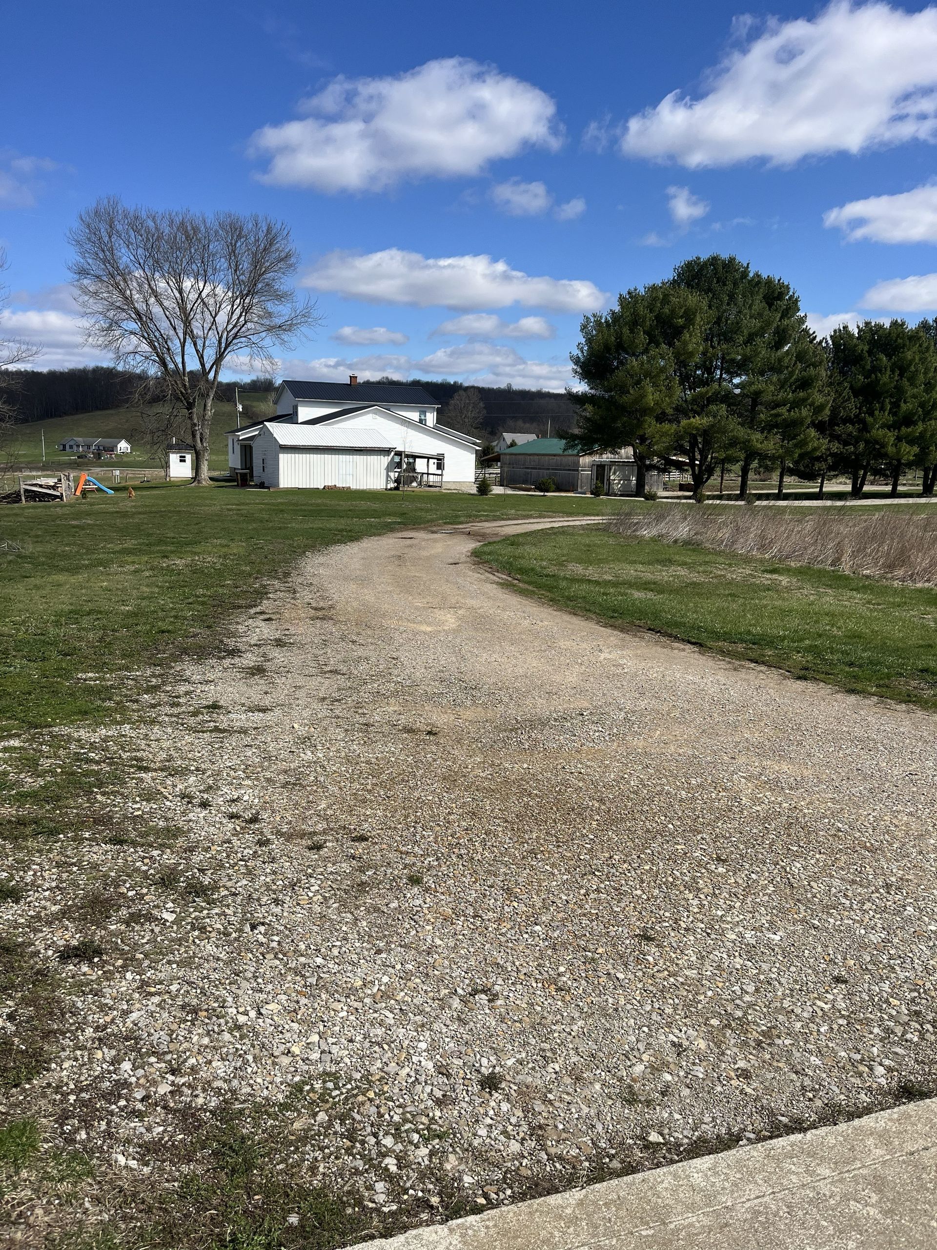 Gravel path leading to a white farmhouse under a blue sky with clouds, flanked by trees and grassy fields
