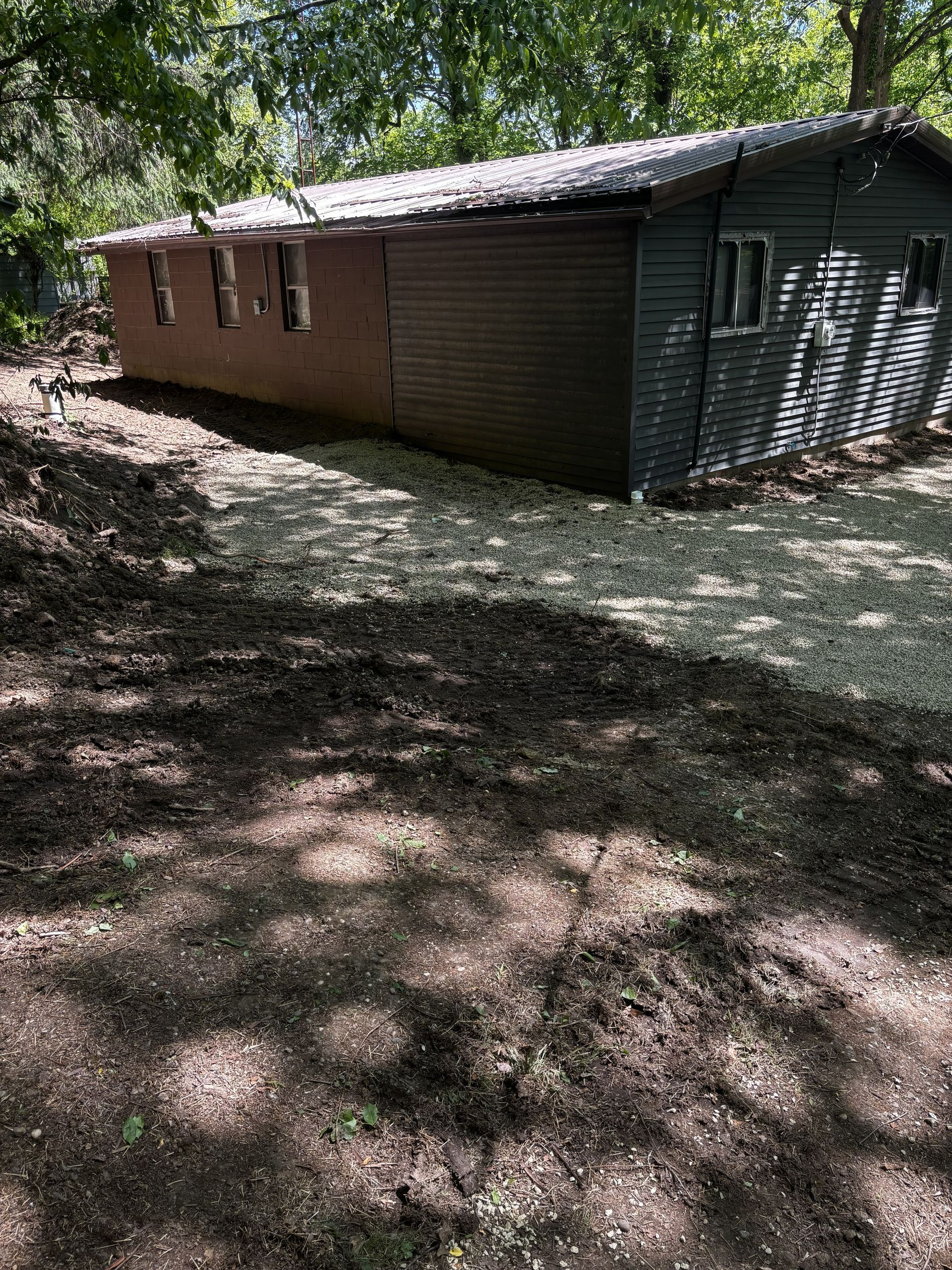Brick building in a wooded area with a corrugated metal roof and shaded dirt ground