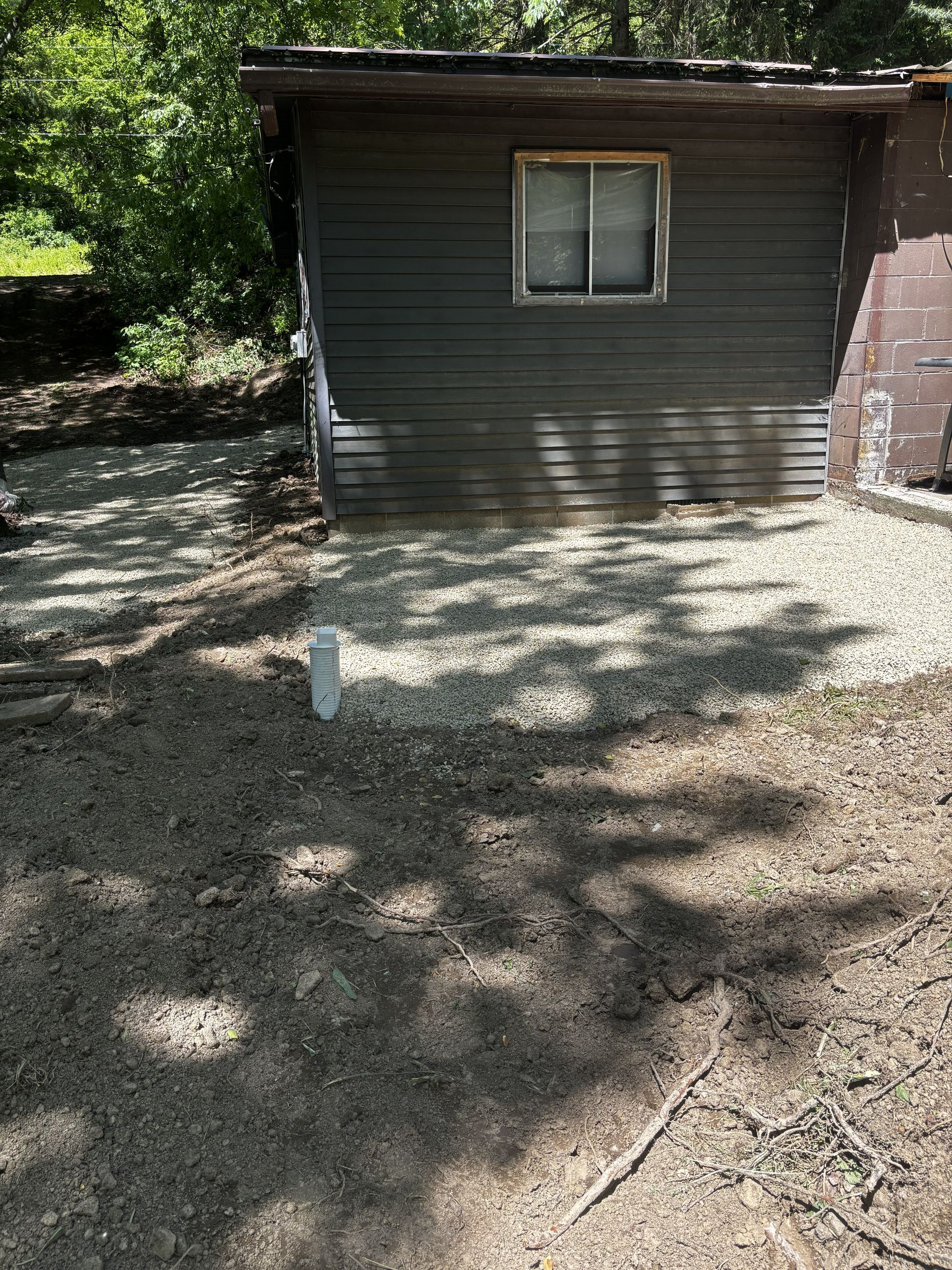 Small brown shed with a window in a shaded gravel yard, with trees in the background.