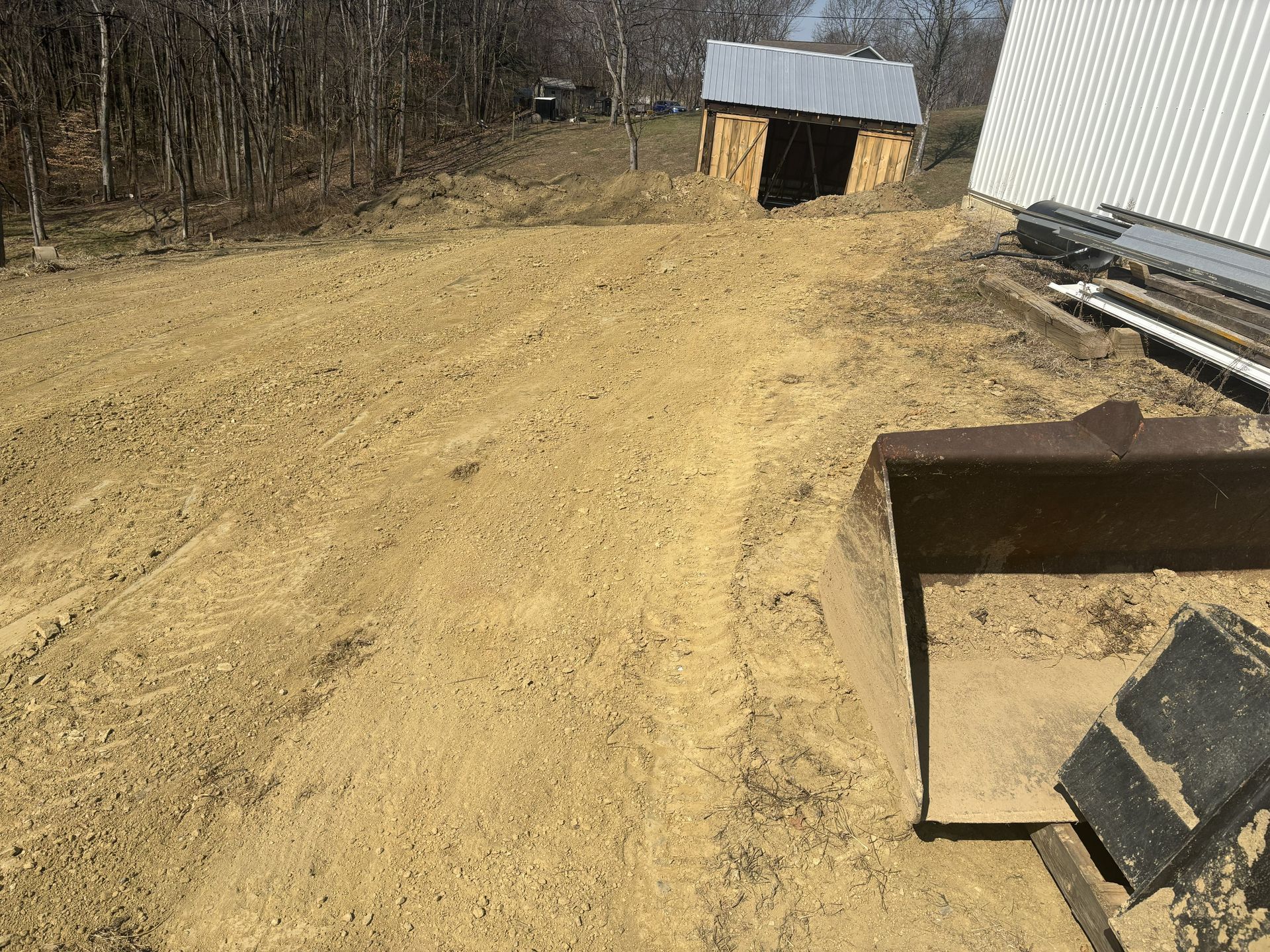 Dirt path beside a barn and shed, with a metal dumpster and stacked pipes on the right.