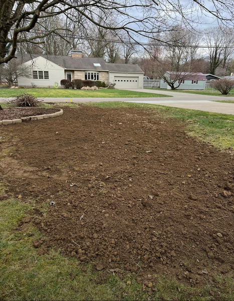 Front yard with bare soil, a small landscaped bed, and suburban houses in the background.