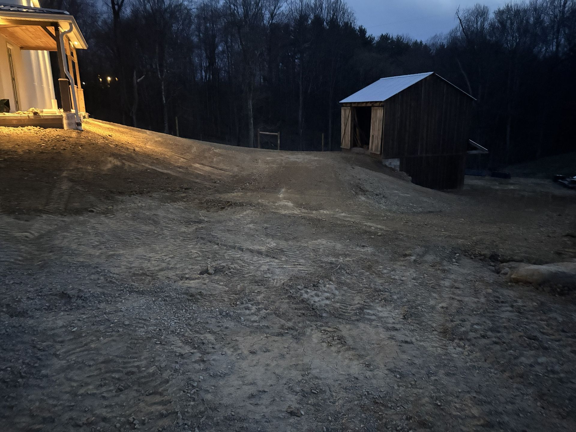 Dim gravel yard at dusk with a lit house on the left and a small shed near a dark treeline