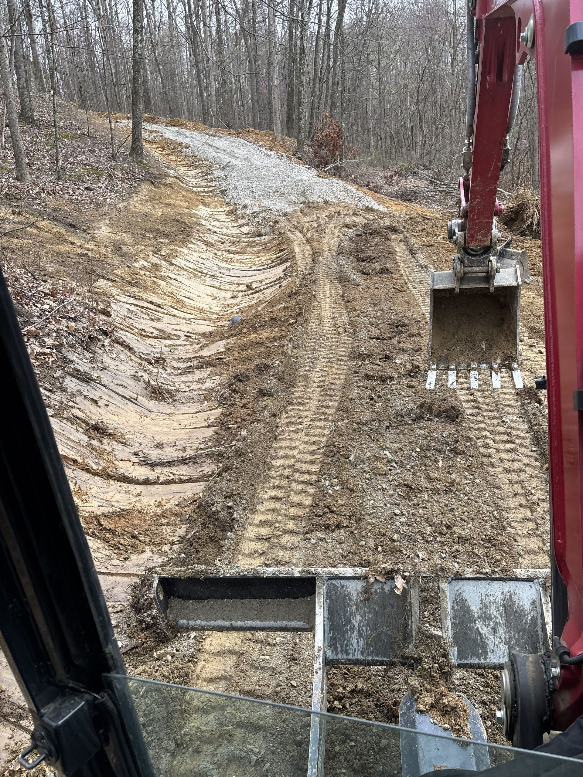 View from excavator cab of muddy dirt road through leafless woods, with bucket visible on the right