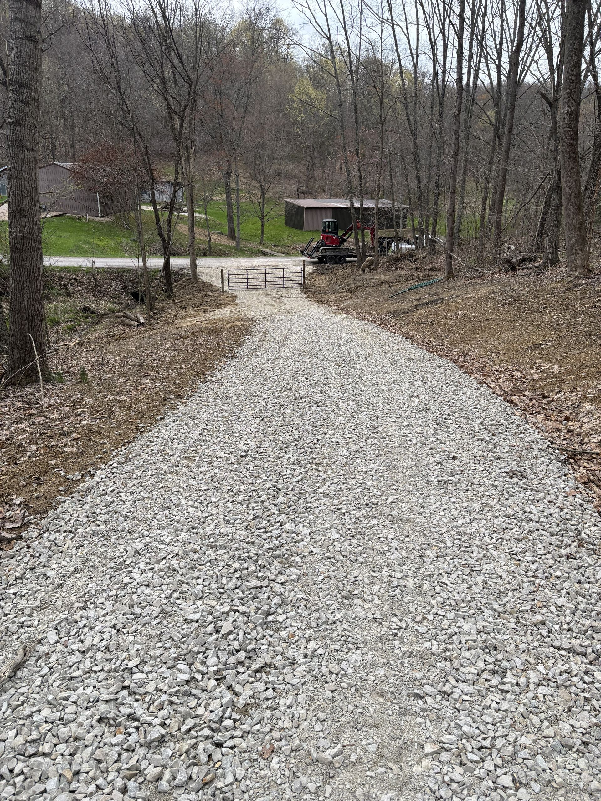 Gravel path through woods leading to a road with a small red tractor in the distance