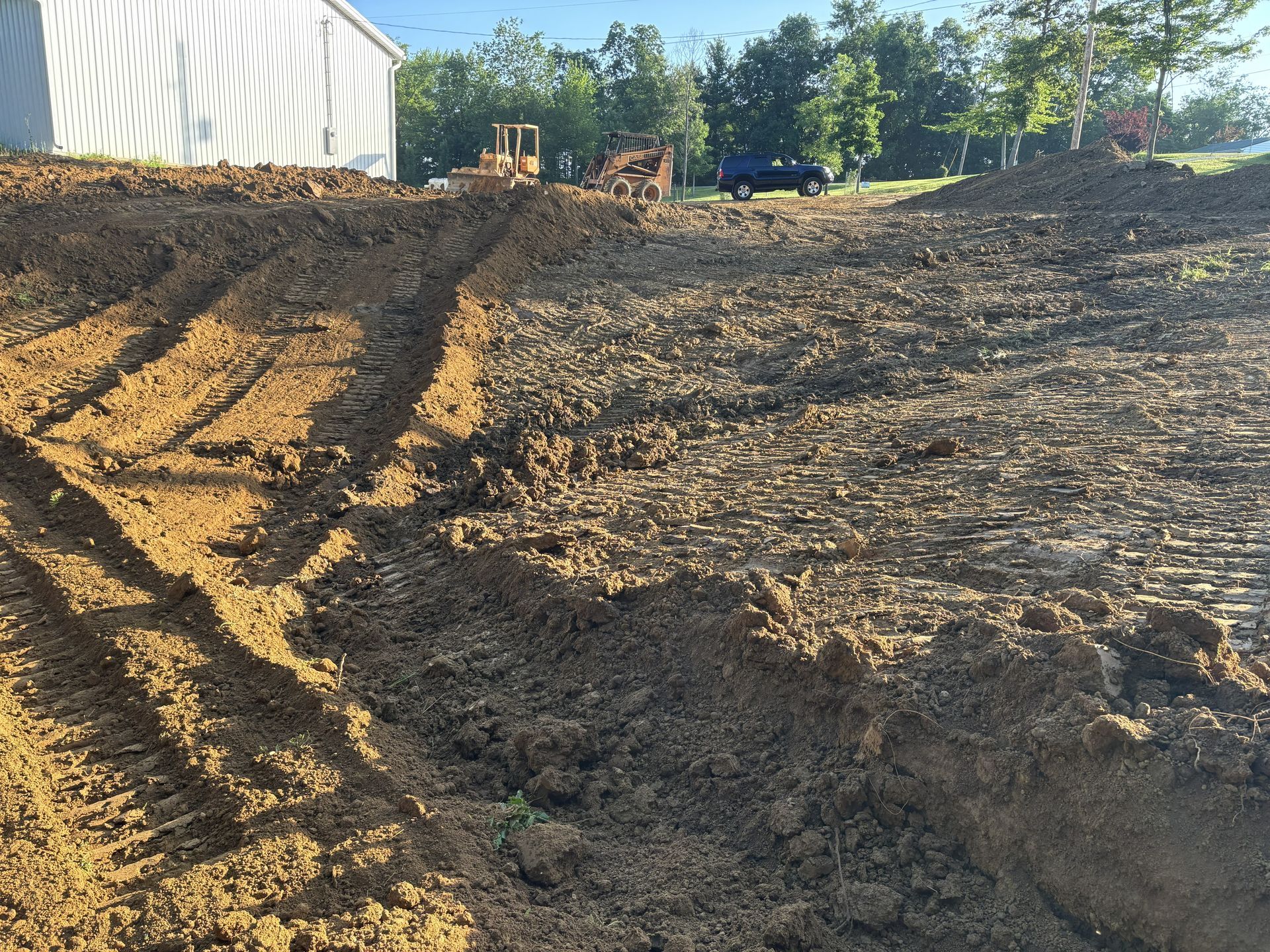 Freshly graded dirt lot beside a white building, with tire tracks and trees in the background