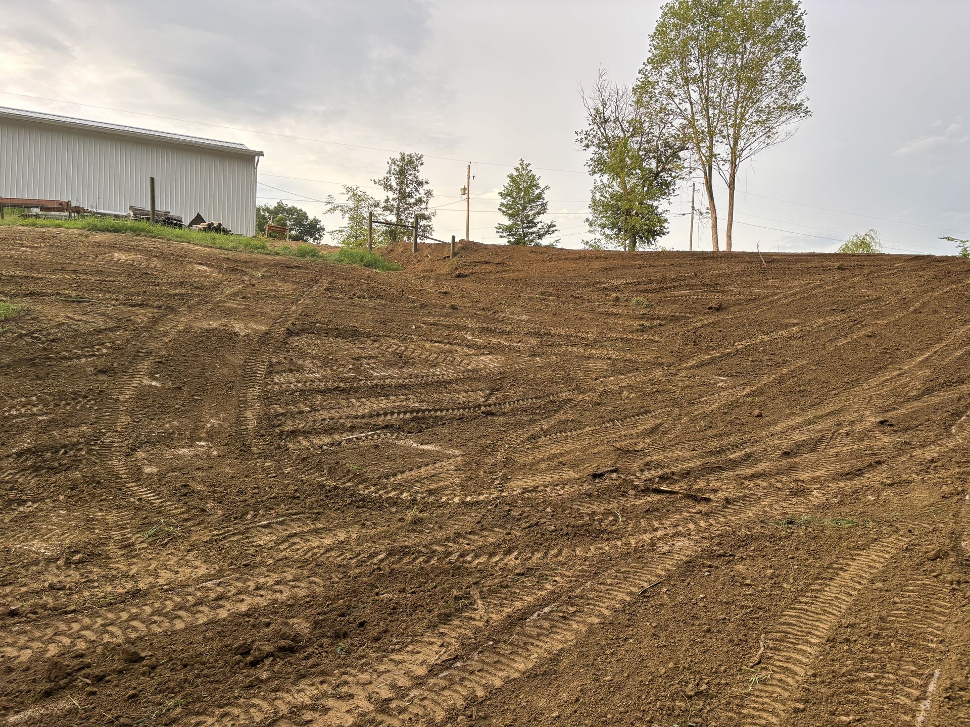 Recently cleared dirt slope beside a white metal building and a few trees under an overcast sky
