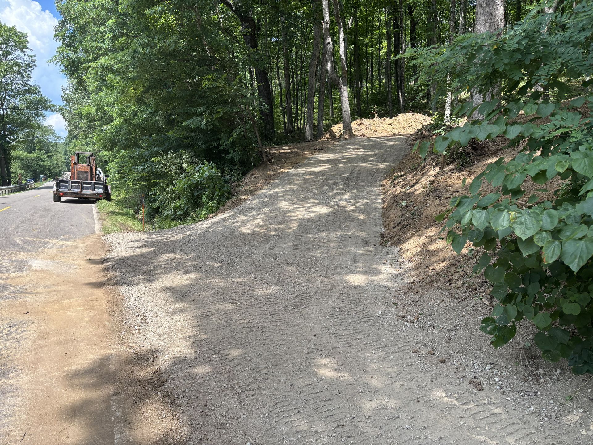 Gravel forest trail curving uphill beside trees, with a vehicle parked on the roadside.