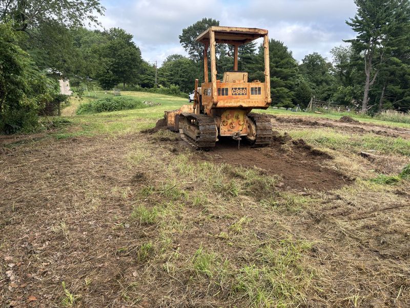 Yellow bulldozer clearing muddy ground in a grassy field with trees in the background