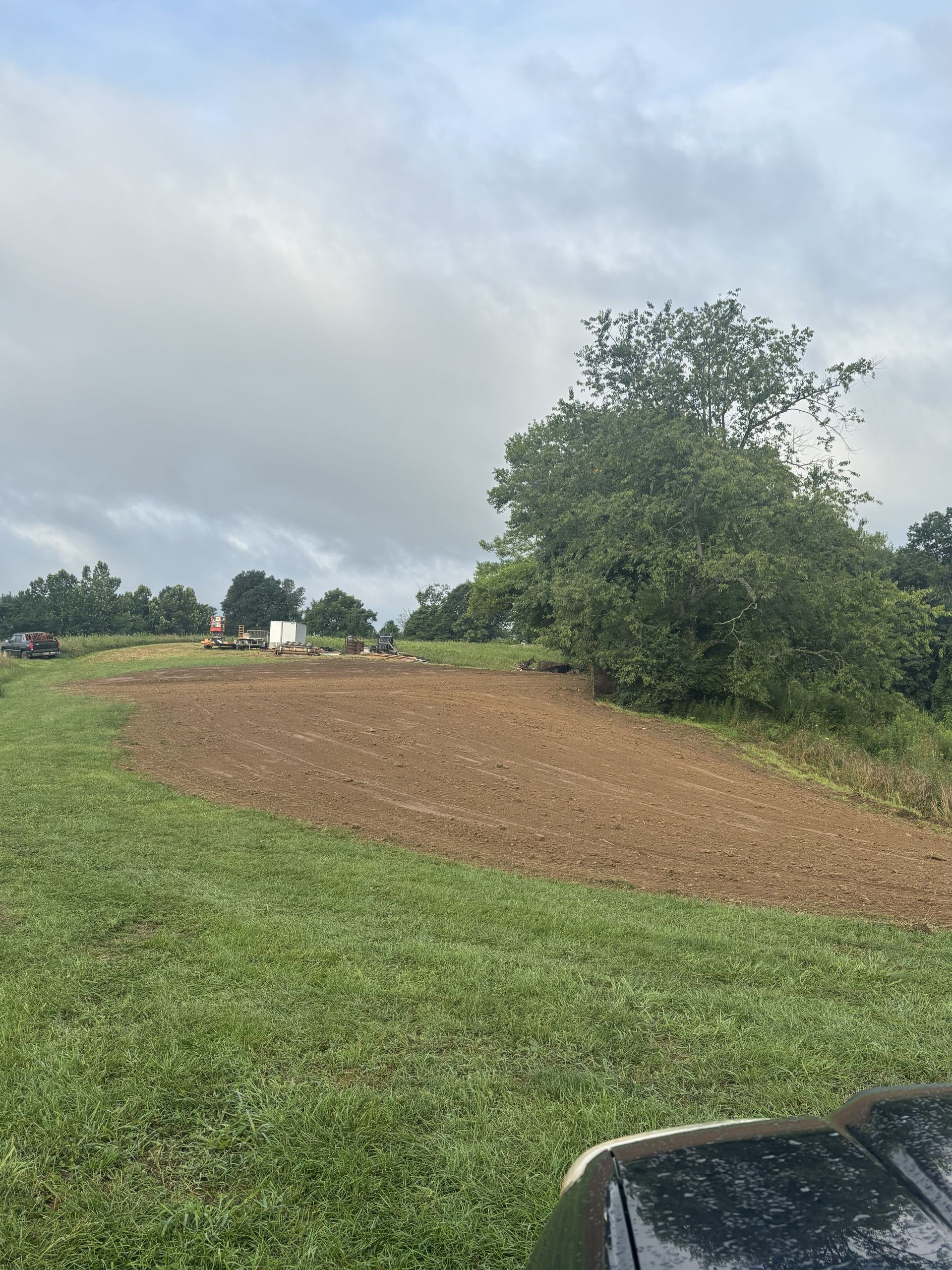 Rural field with brown soil, green grass, a large tree, cloudy sky, and part of a car hood in the foreground
