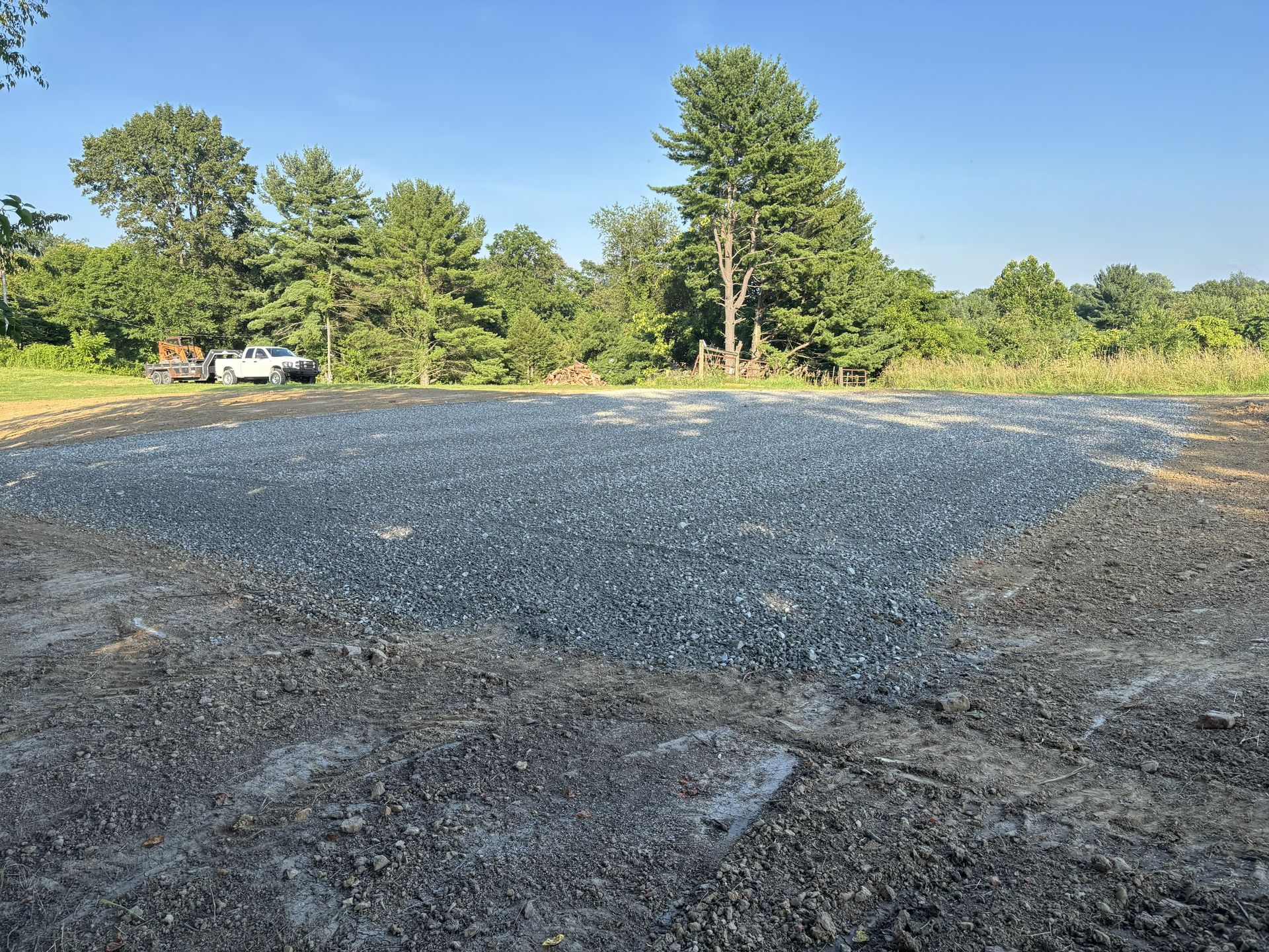 Freshly paved gravel lot under a clear blue sky, bordered by trees and brush