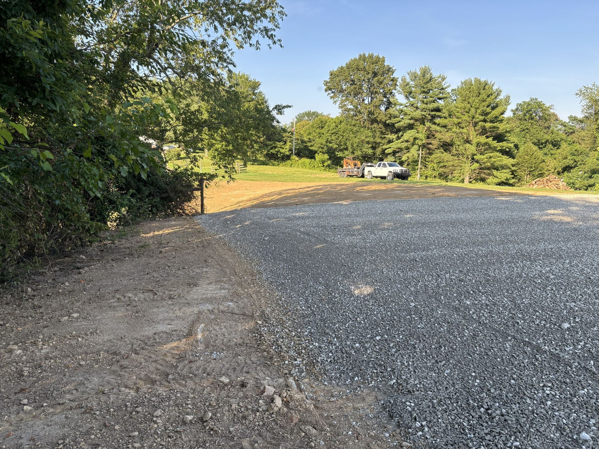 Gravel parking area beside trees and brush, with a small white vehicle in the distance