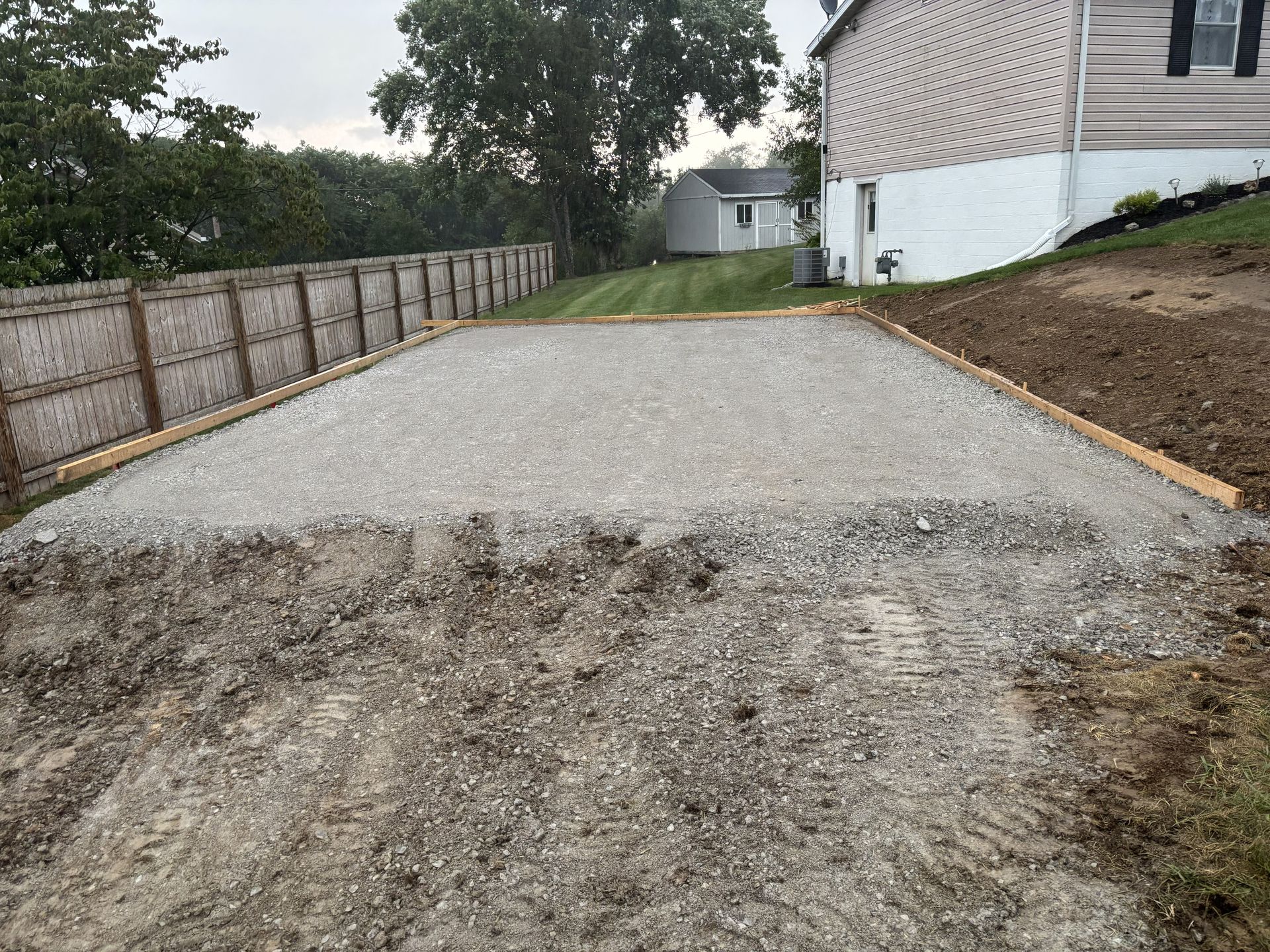 New concrete driveway beside a house, with a rough gravel edge and fenced yard on the left.