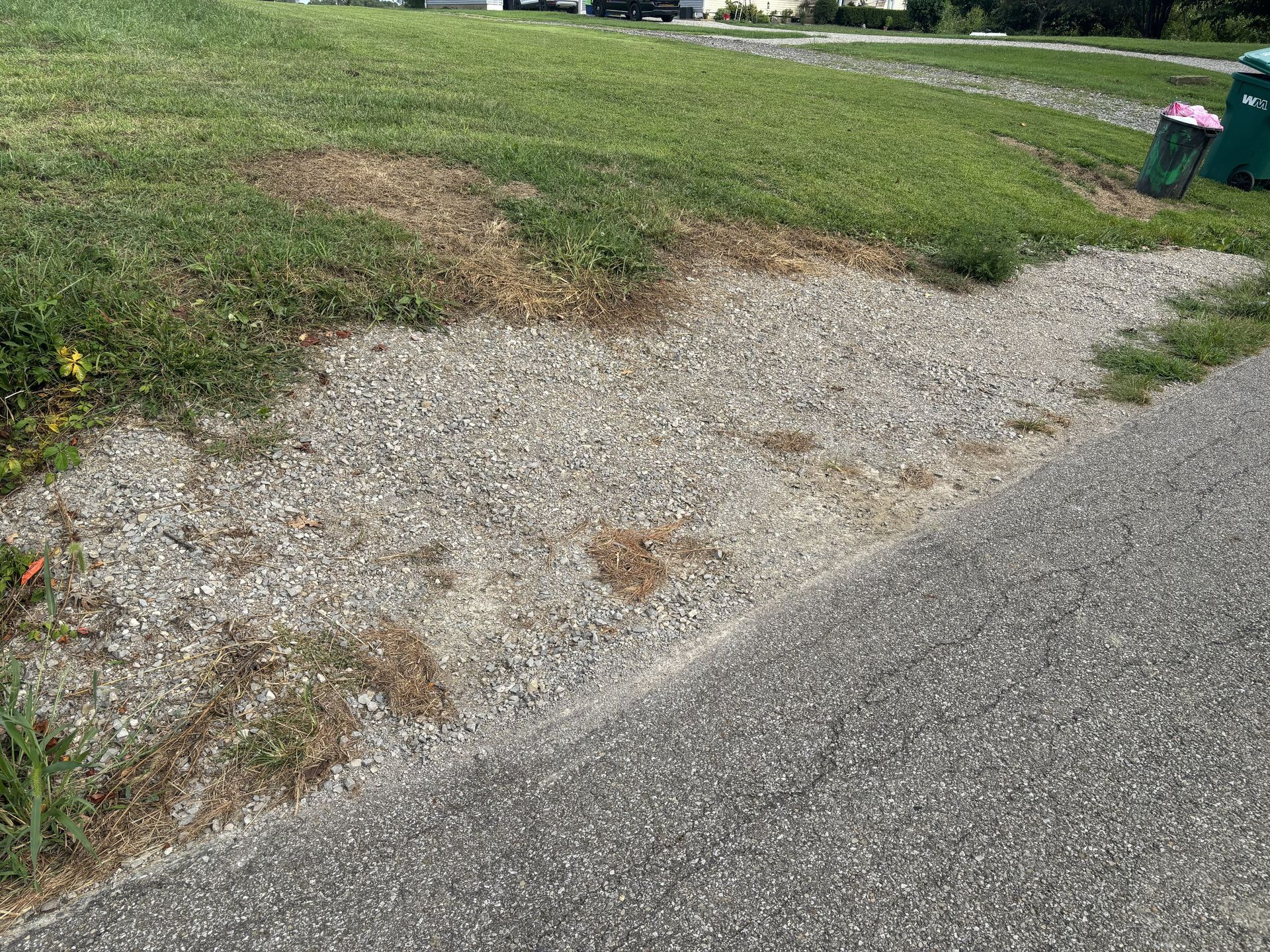 Gravel driveway edge beside grass, with a small patch of bare ground and a trash bin in the distance