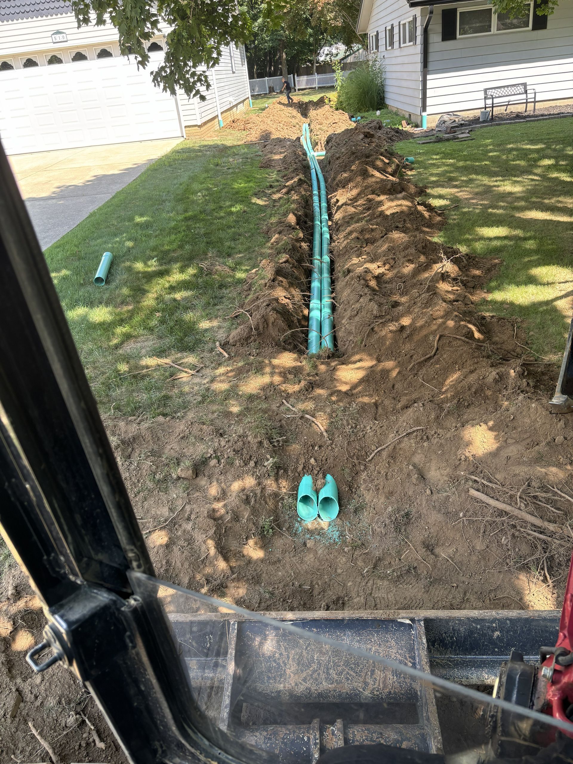 Trench in front yard with teal PVC pipes laid along it beside a white house and driveway.