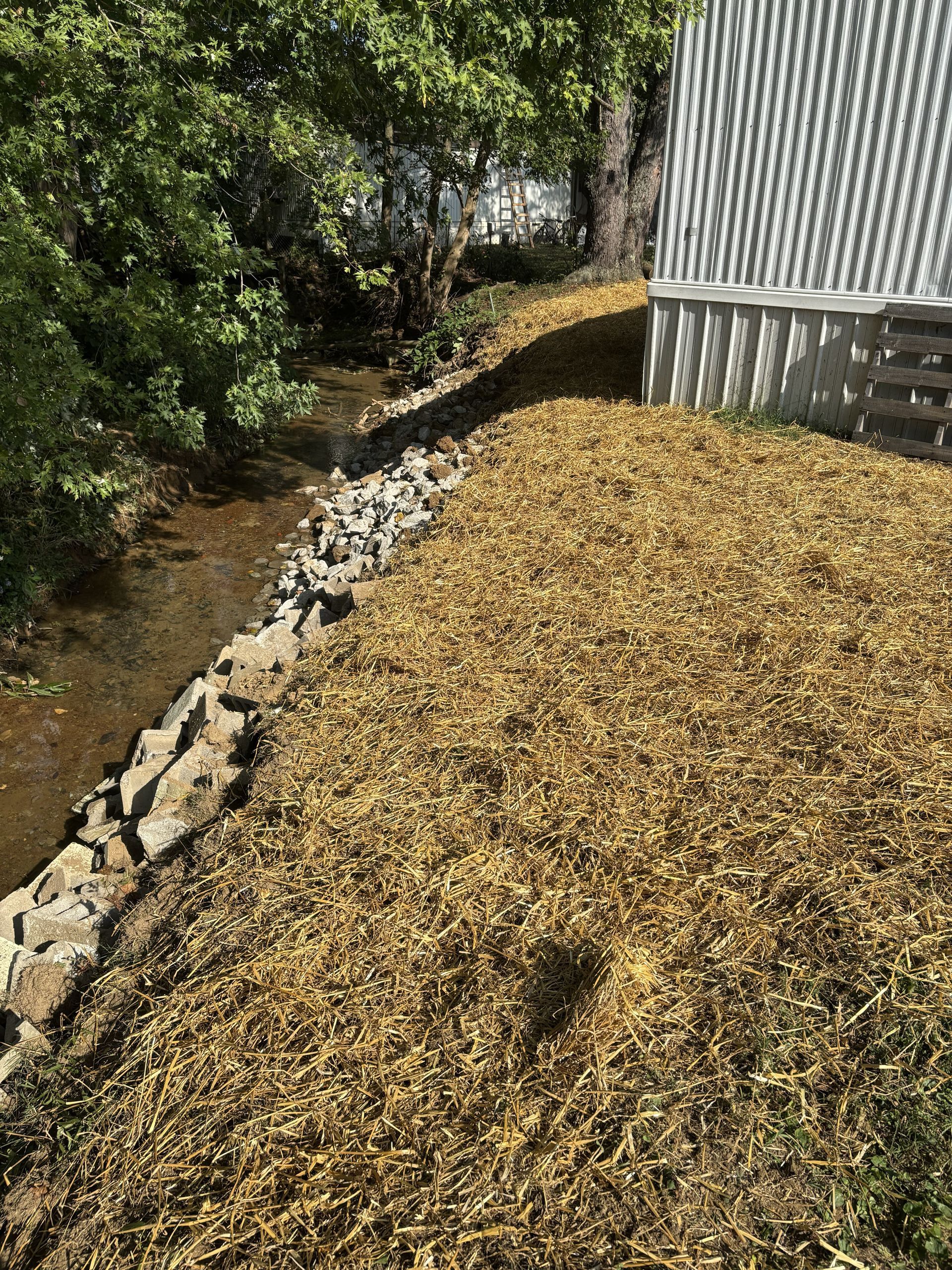 Mulched slope beside a stream and a corrugated metal building, with rocks lining the bank.