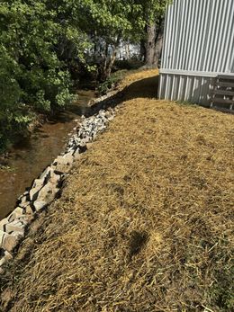 Mulch-covered slope beside a small stream and a corrugated metal building, with rocks lining the bank