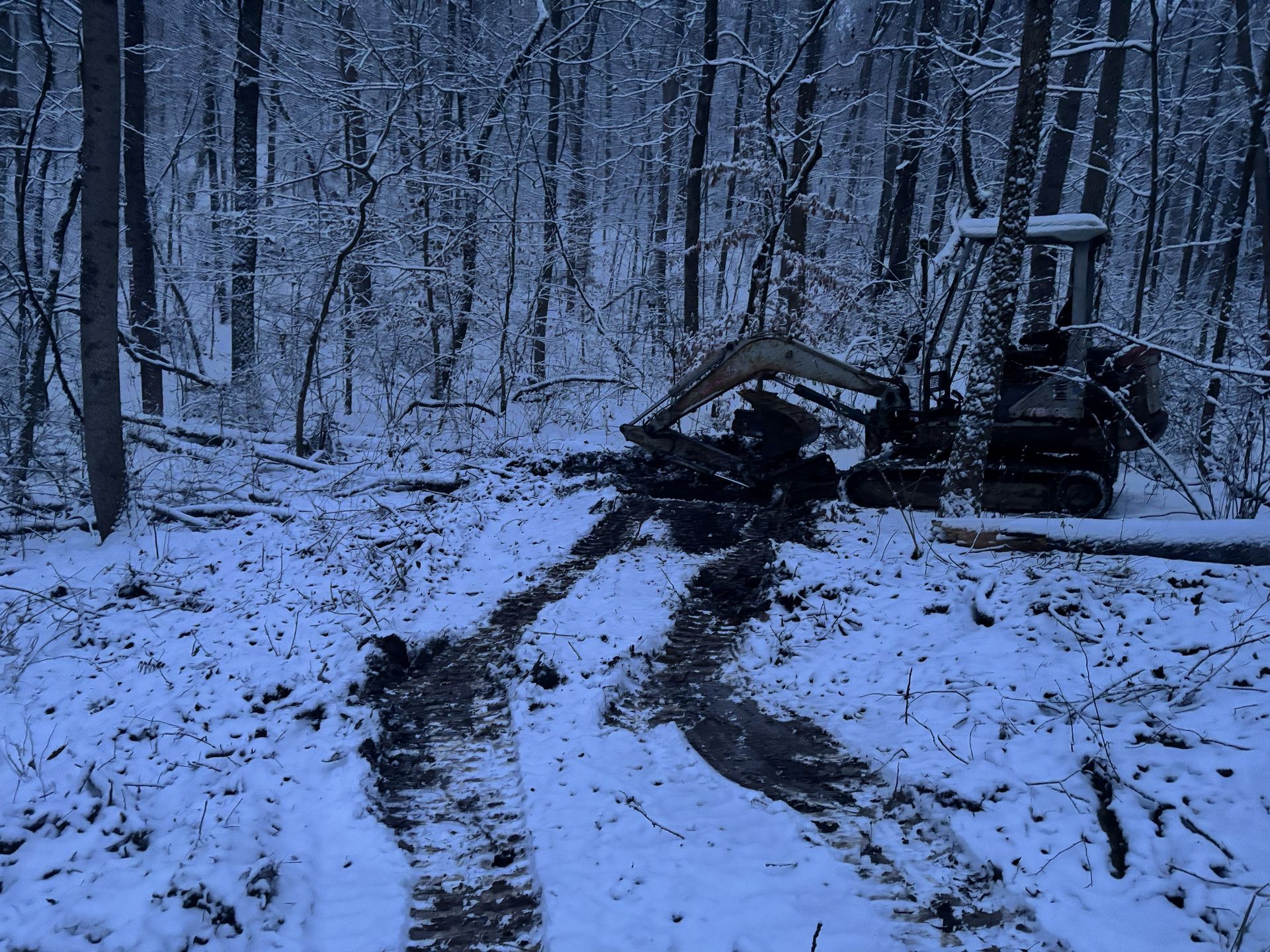 Snowy forest trail with tire tracks and a small excavator clearing debris at the path edge