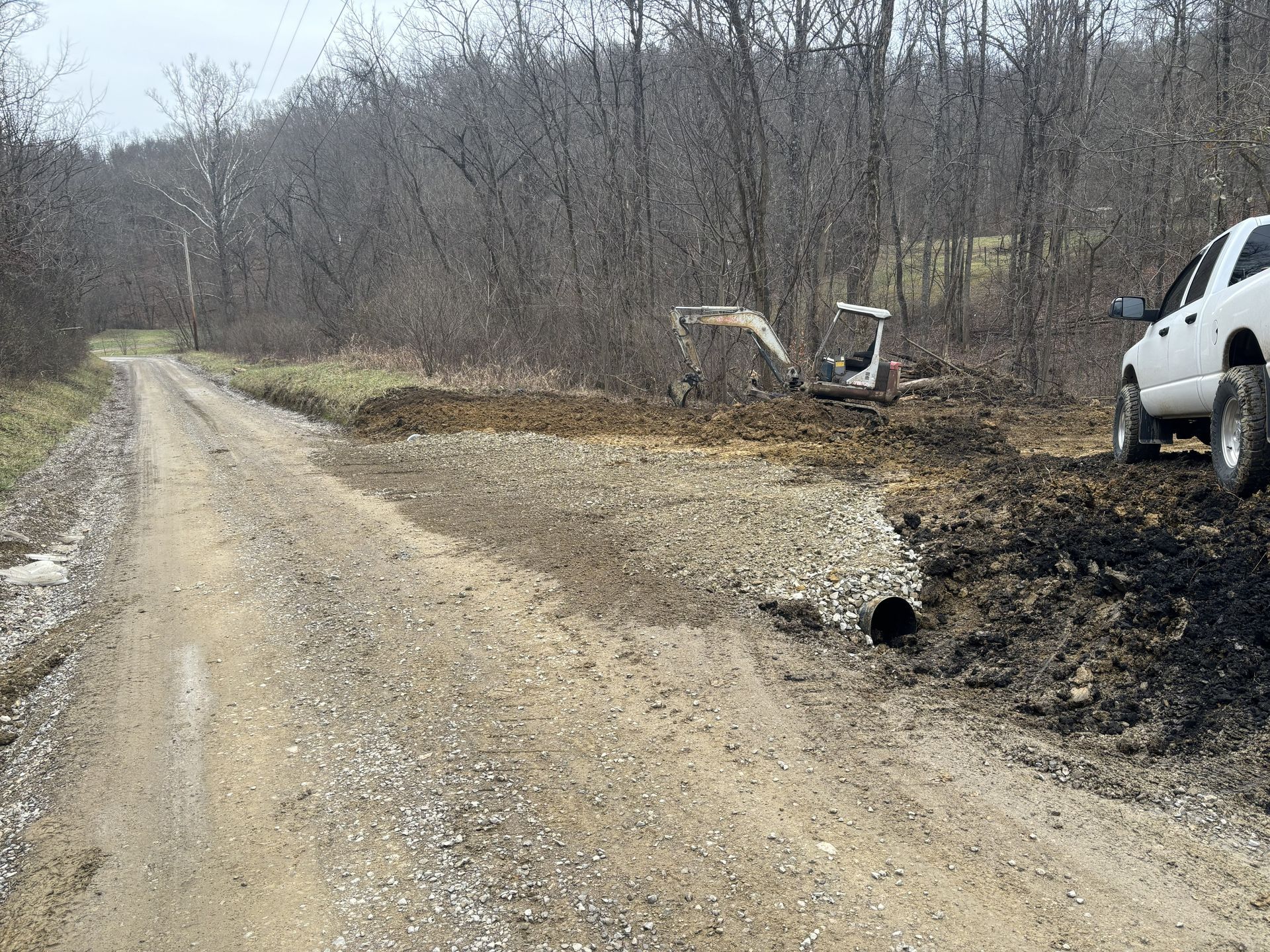 Gravel road through bare woods with a white truck at right and a small excavator near a burned area.