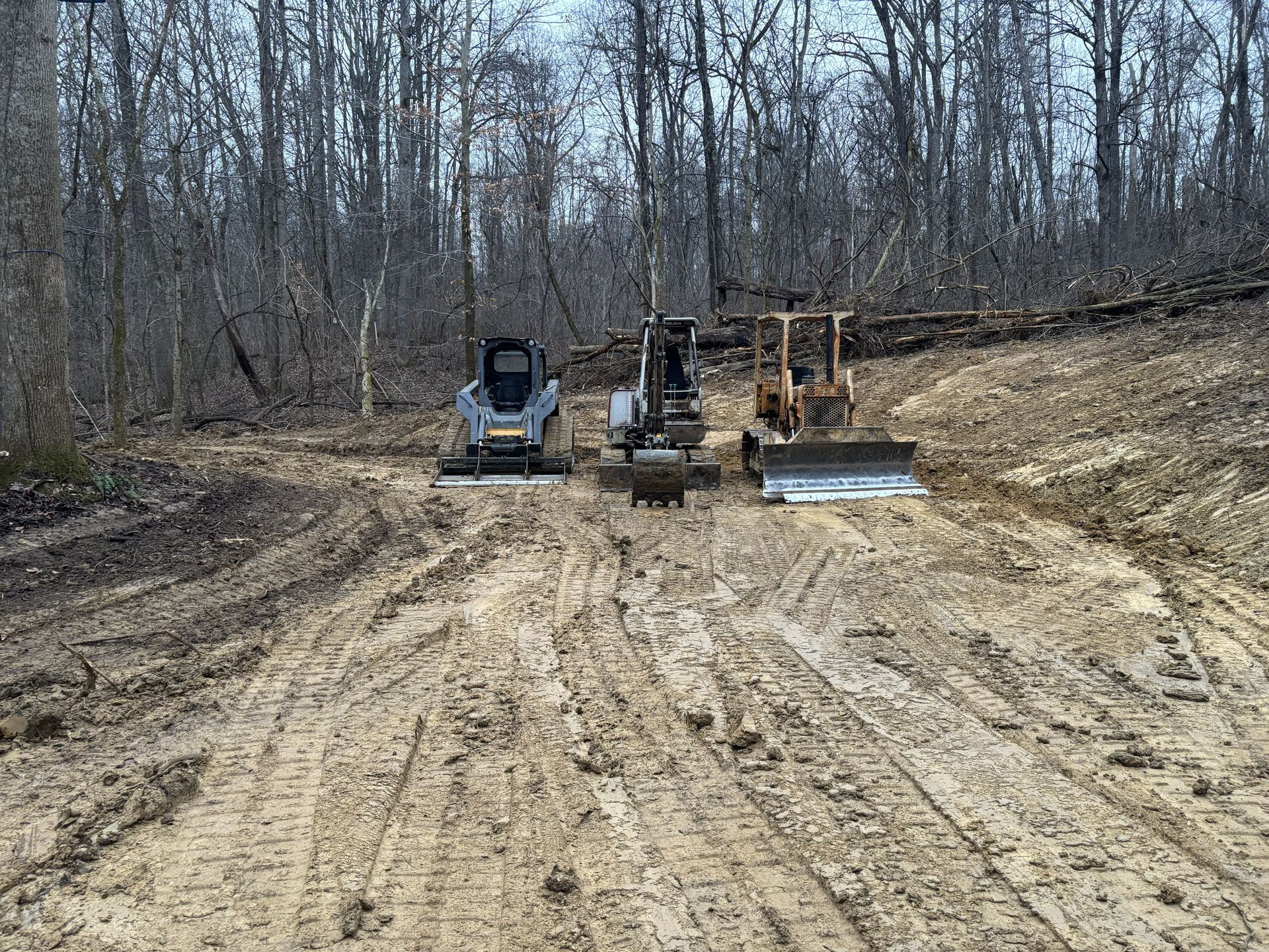 Three construction vehicles on a muddy dirt road in a leafless wooded area
